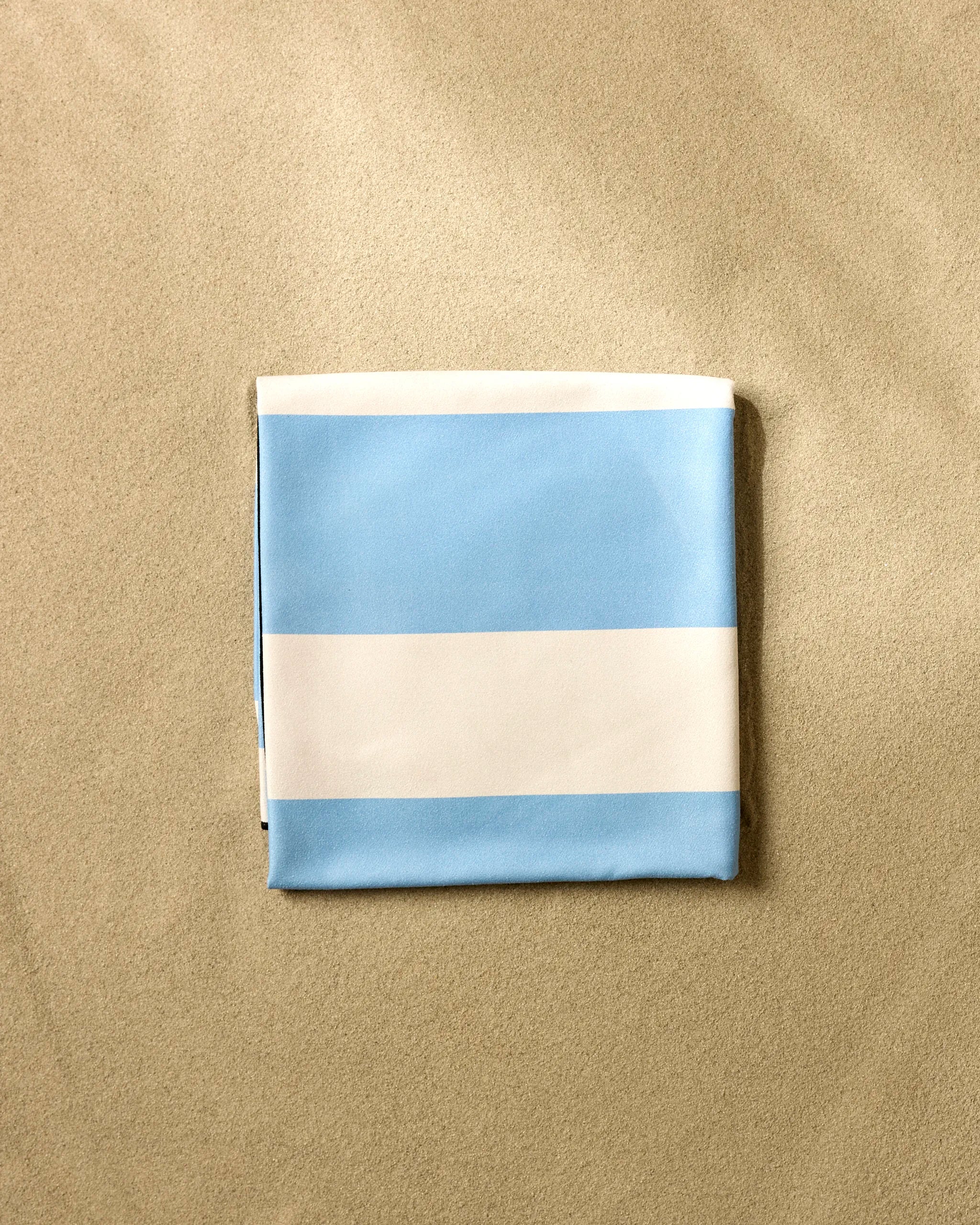 A GEOMETRY Blue Block Stripes Micro-loop Beach Blanket with alternating light blue and white stripes is neatly folded on the sand.