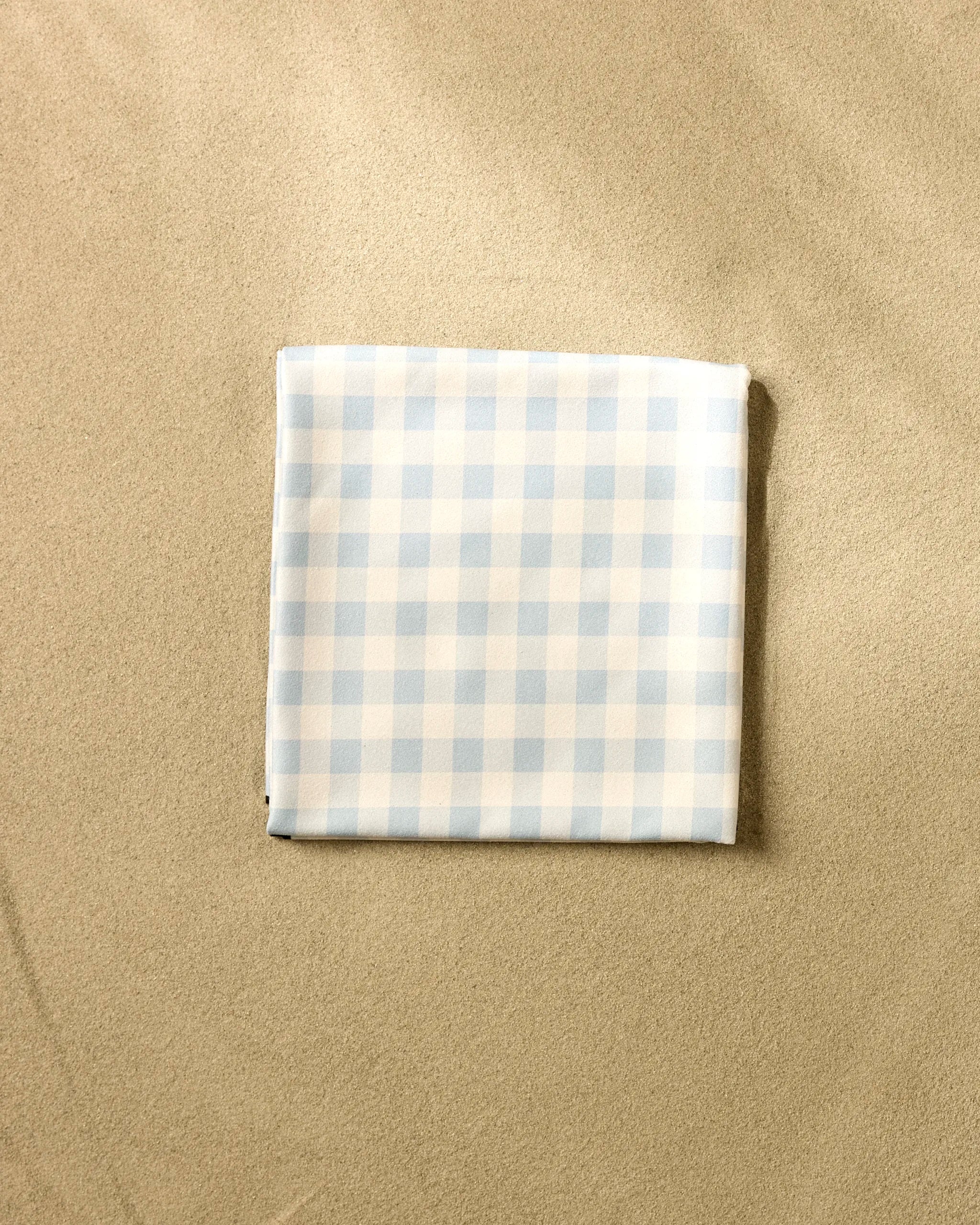The Aubrey Fairchild Blue Gingham beach blanket, crafted from quick-drying recycled materials, is folded on smooth beige sand and displays a light blue and white checkered pattern.