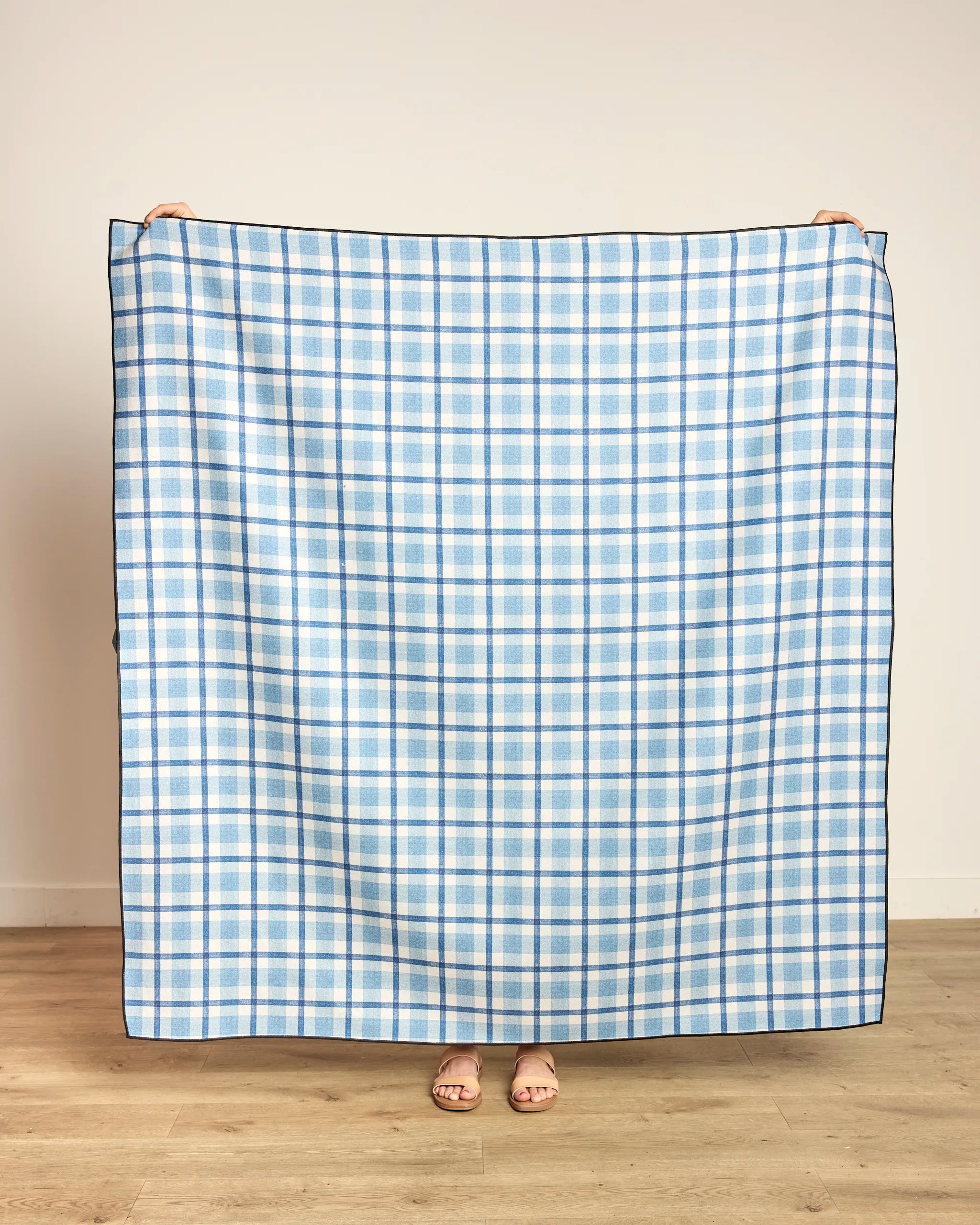 A person stands on a wooden floor holding up the Geometry Bluefield recycled beach blanket in blue and white plaid, which covers their body except for hands and feet.