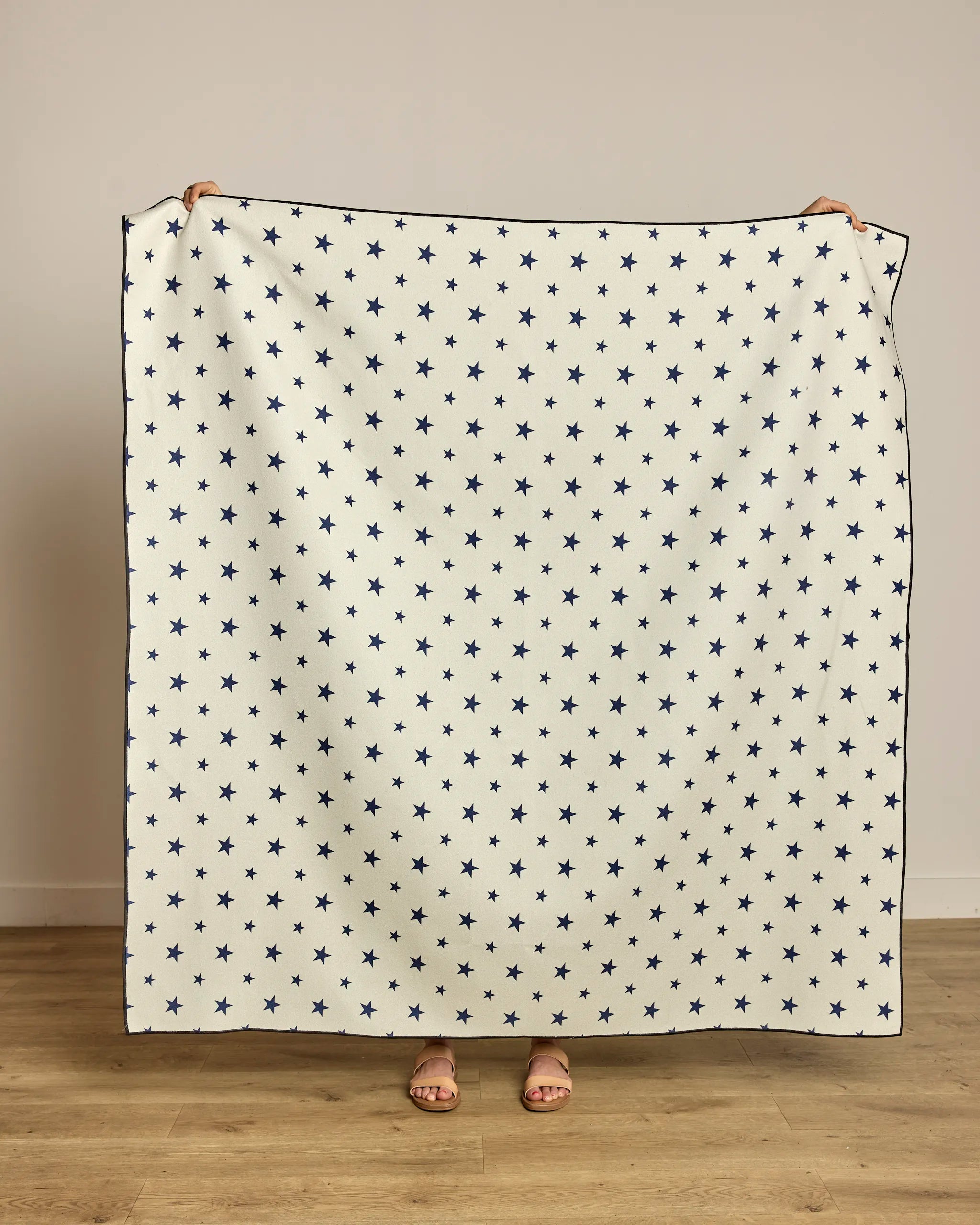 A person holds up the Geometry Boardwalk Star, a large, quick-drying white beach blanket with a blue star pattern, covering their body and face. Only their hands and sandal-clad feet are visible against a plain wall and wooden floor.