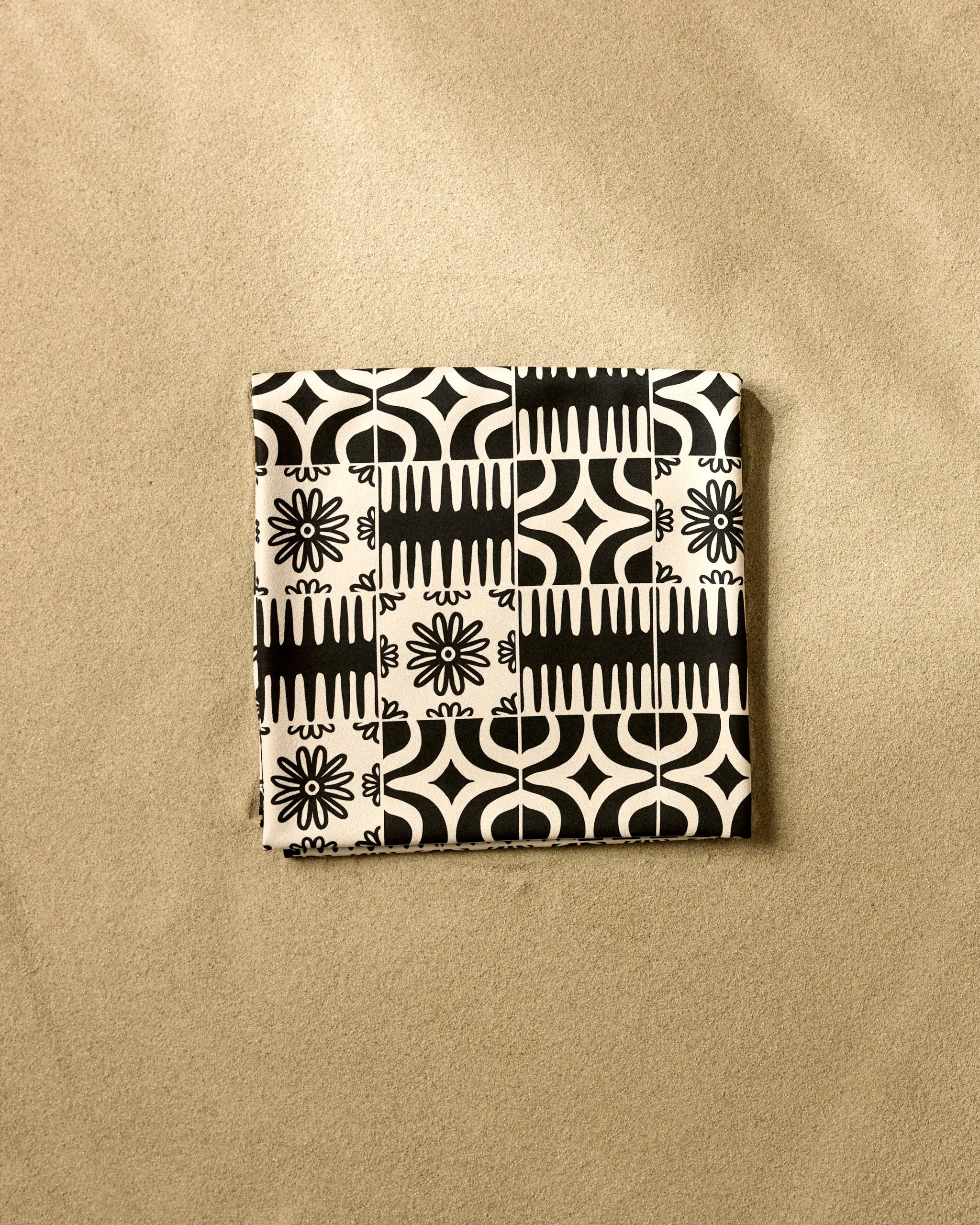 The GEOMETRY Bohemian Mosaic is a square recycled beach blanket featuring a black and white geometric and floral pattern, displayed on a textured beige surface.