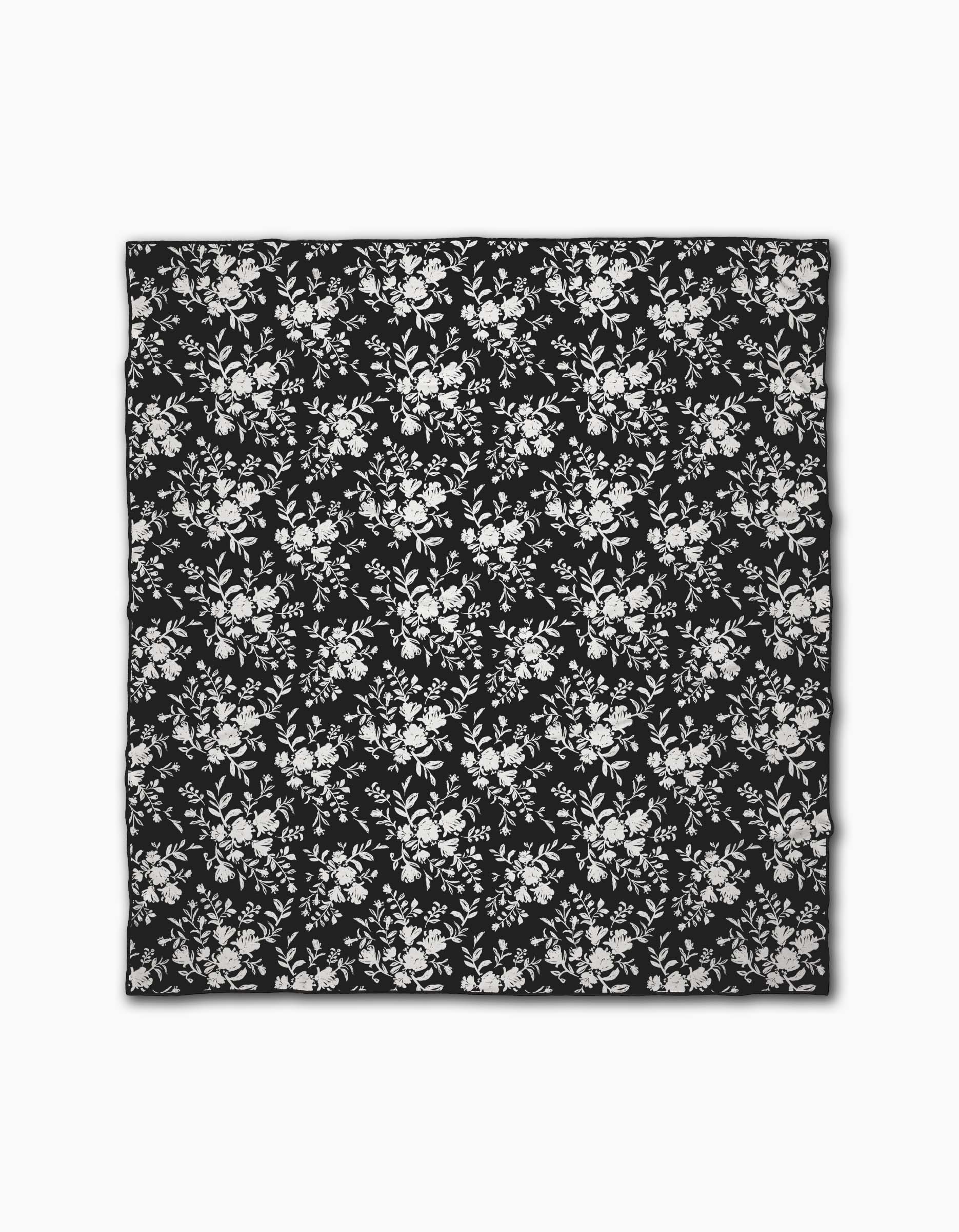 The Danelys Sidron Botanical Silhouettes Charcoal is a black square beach blanket made from quick-drying, recycled materials, showcasing a white floral pattern with evenly spaced clusters of flowers and leaves.