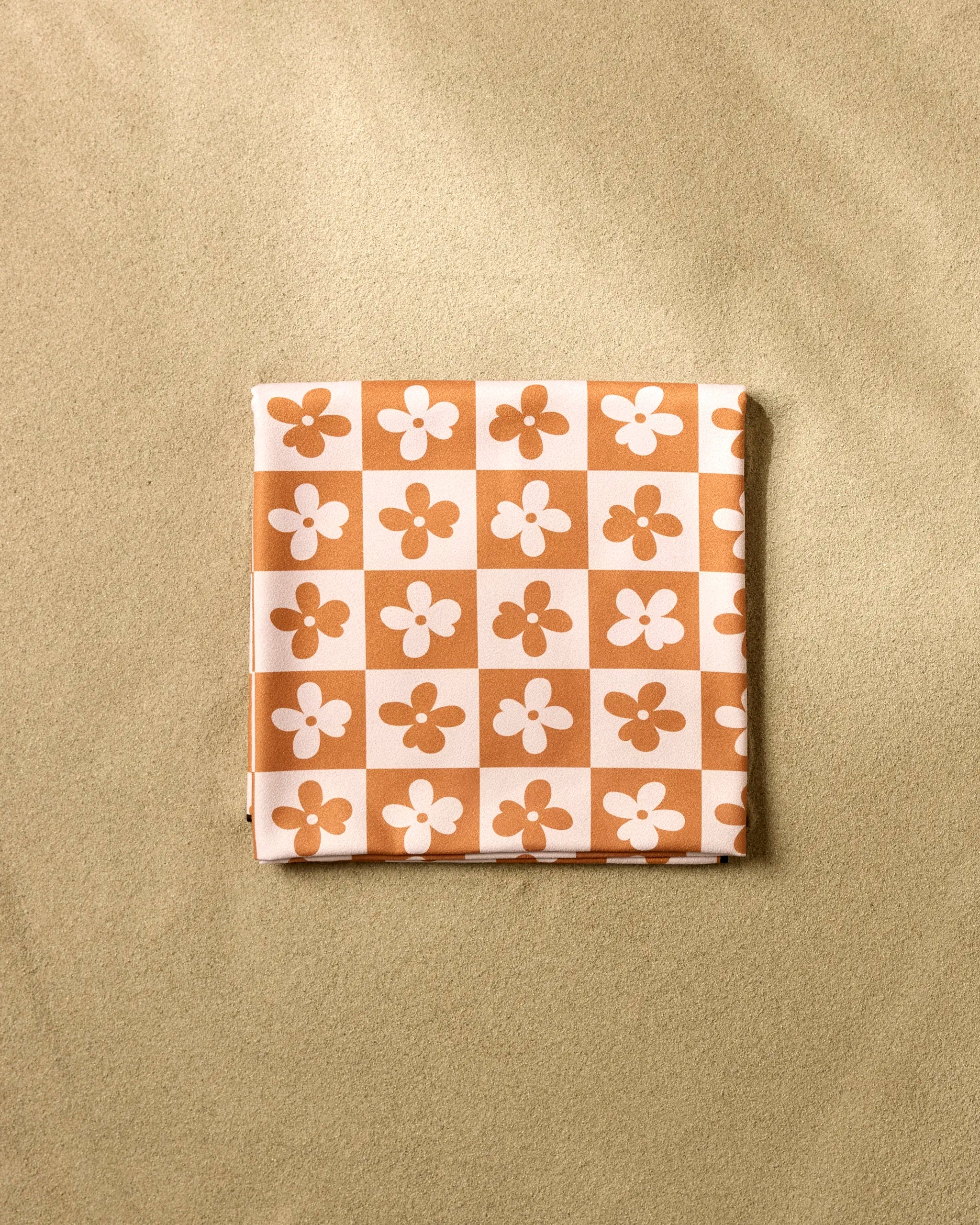 The GEOMETRY Burnt Orange Flowers beach blanket, made from quick-drying recycled materials, features a brown and cream checkerboard pattern with white and brown flower designs—ideal for eco-friendly relaxation on the sand.