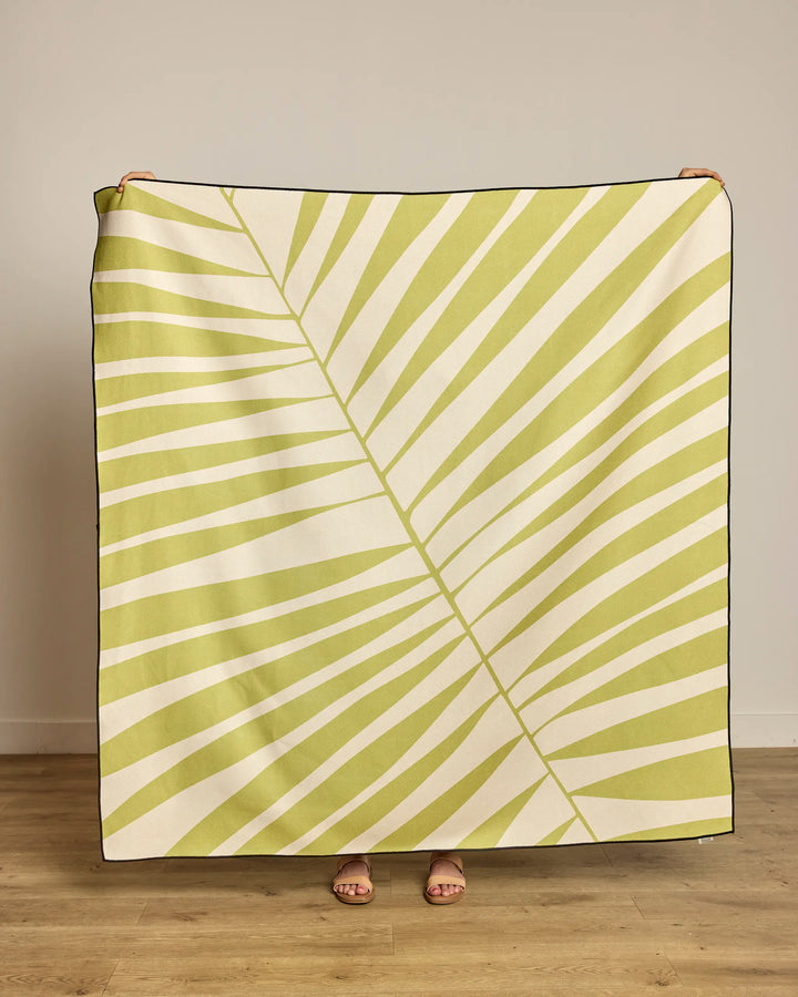 A person stands behind the Geometry Cabana Club beach blanket, featuring a light green palm leaf design on cream. Made from recycled materials, only their hands and feet show against a plain wall and wooden floor.