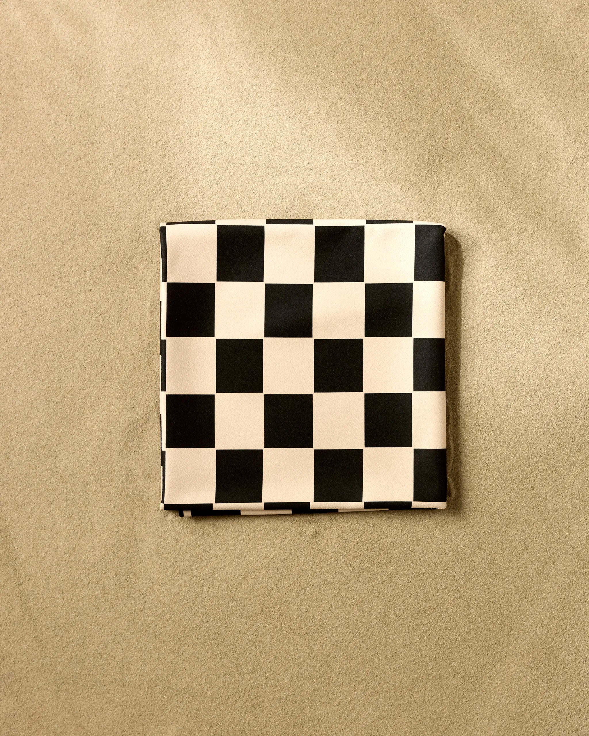 The GEOMETRY Classic Checker beach blanket, quick-drying and checkered in black and white, is neatly folded into a rectangle on smooth, light sand.
