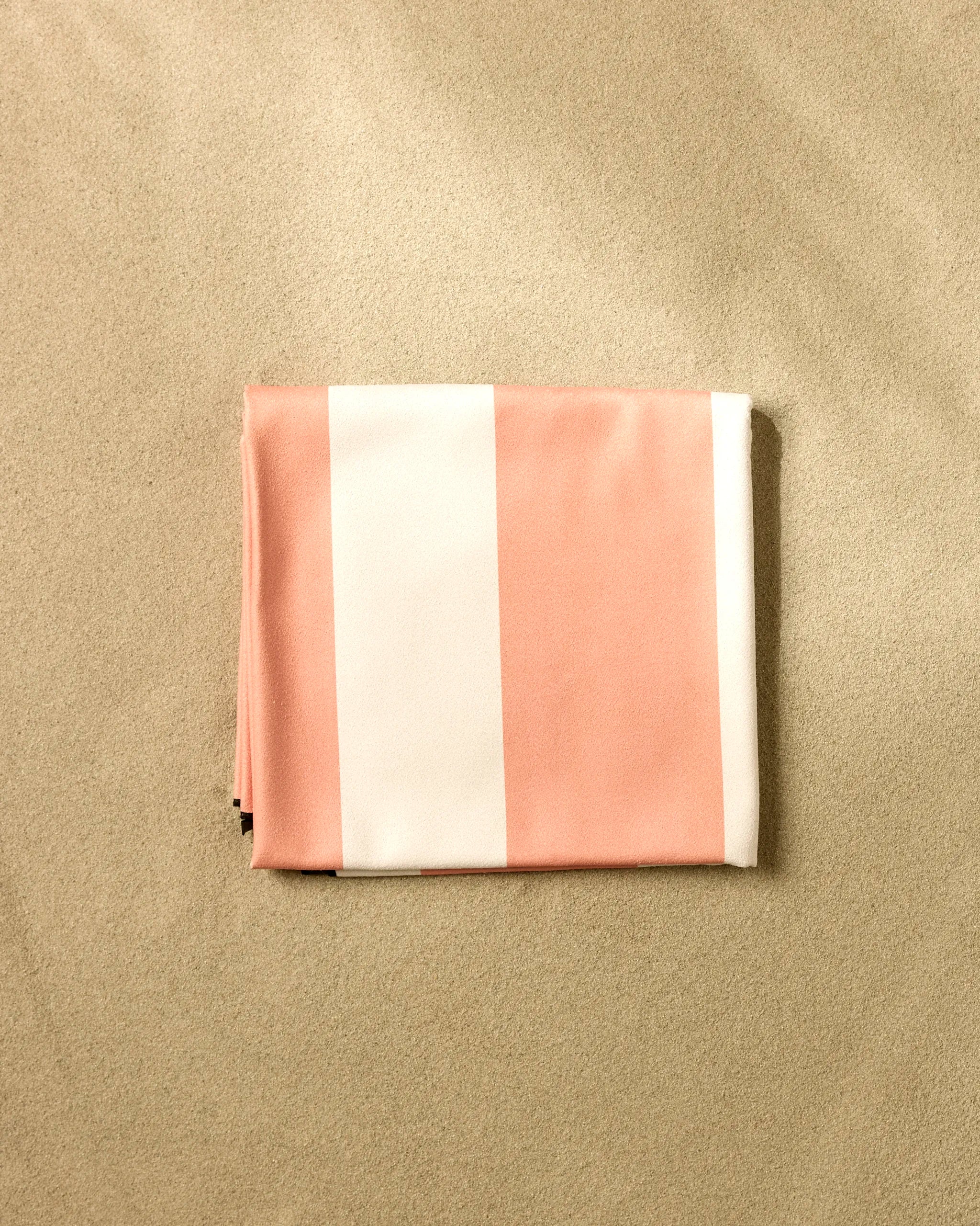 The GEOMETRY Coral Block Stripes beach blanket, featuring wide pink and white stripes and crafted from quick-drying recycled materials, rests on smooth light sand.
