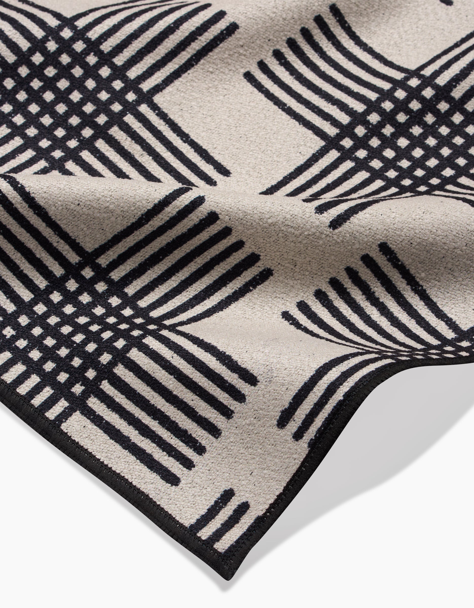 The Cross Hatch Beach Blanket by Rosa Kichline features a beige recycled fabric with a black geometric intersecting line design forming curved squares, shown slightly folded to highlight its textured surface and edge detail.