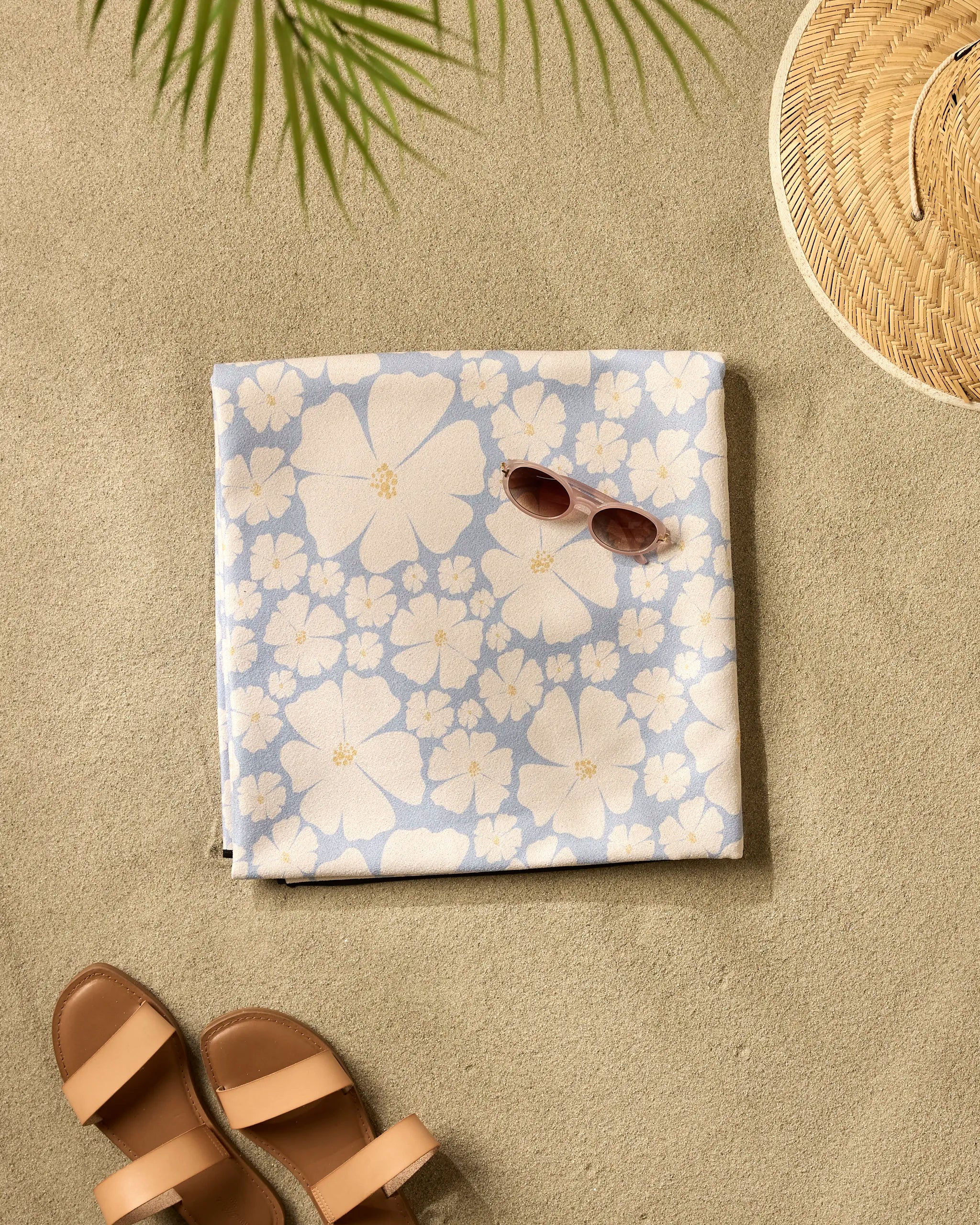 The Daisy Girl Blue beach blanket by Charlie Rowan Designs, crafted from quick-drying recycled materials for eco-friendly comfort, lies on sand with sunglasses, tan sandals, a woven sun hat, and a palm leaf casting shade.