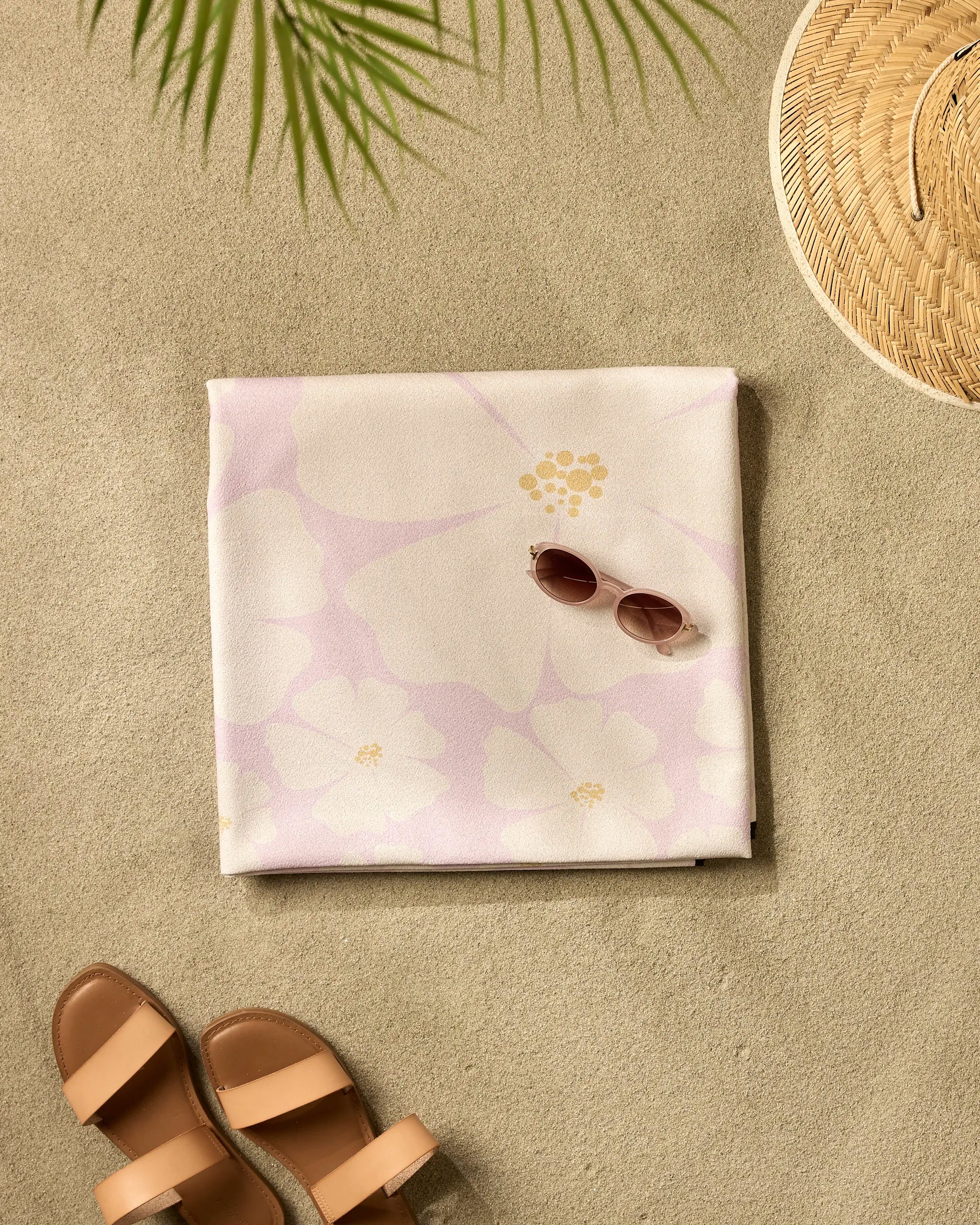 The Daisy Girl Pink beach blanket by Charlie Rowan Designs is displayed on sand with a pink floral towel, sunglasses, tan sandals, a straw hat, and palm leaf shadows—perfect for quick-drying, effortless seaside relaxation.