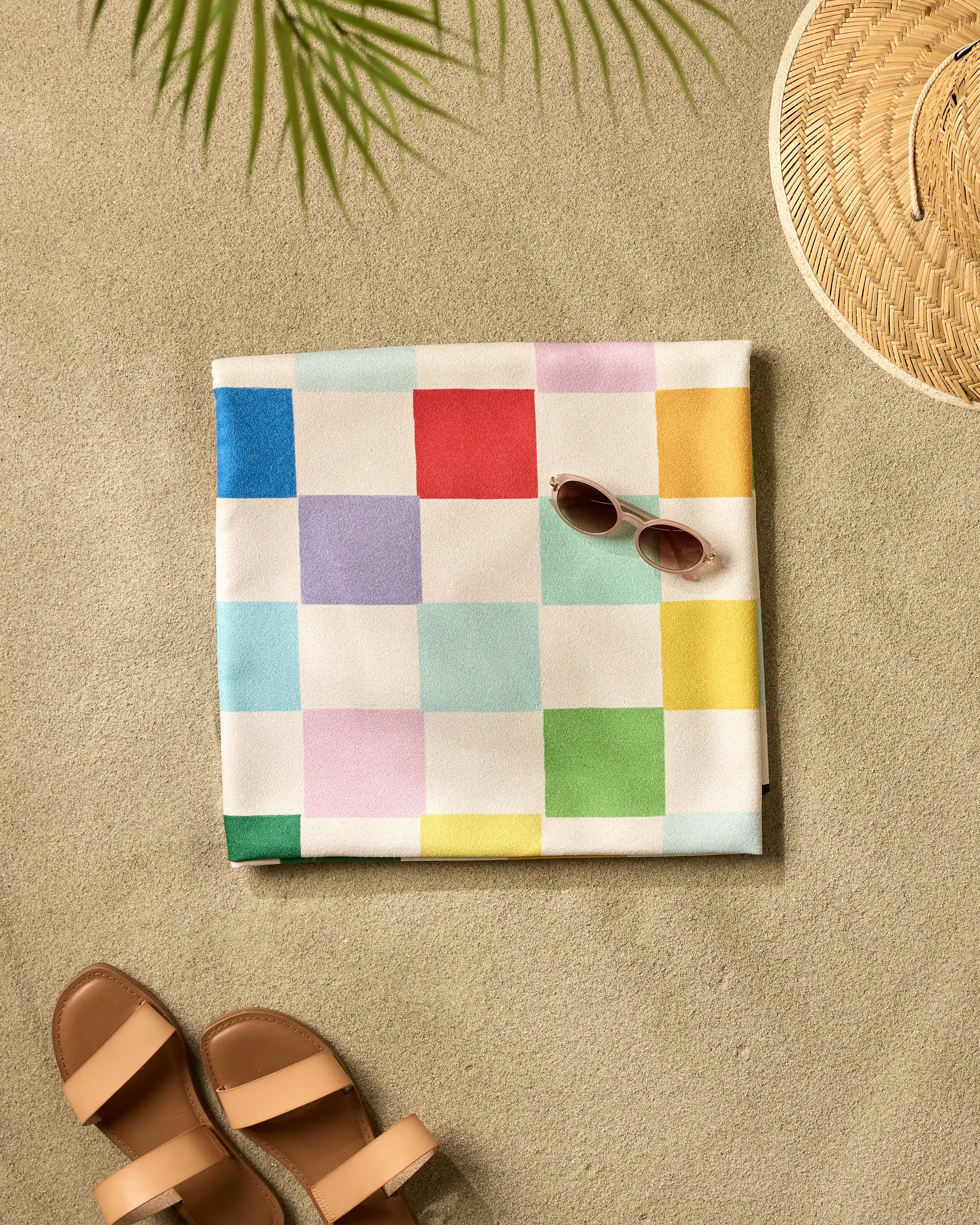 A folded "Dance Floor" beach blanket by Vaishali Design Studio with a colorful checkered pattern lies on the sand beside brown sandals, sunglasses, a woven hat, and a palm leaf casting its shadow.
