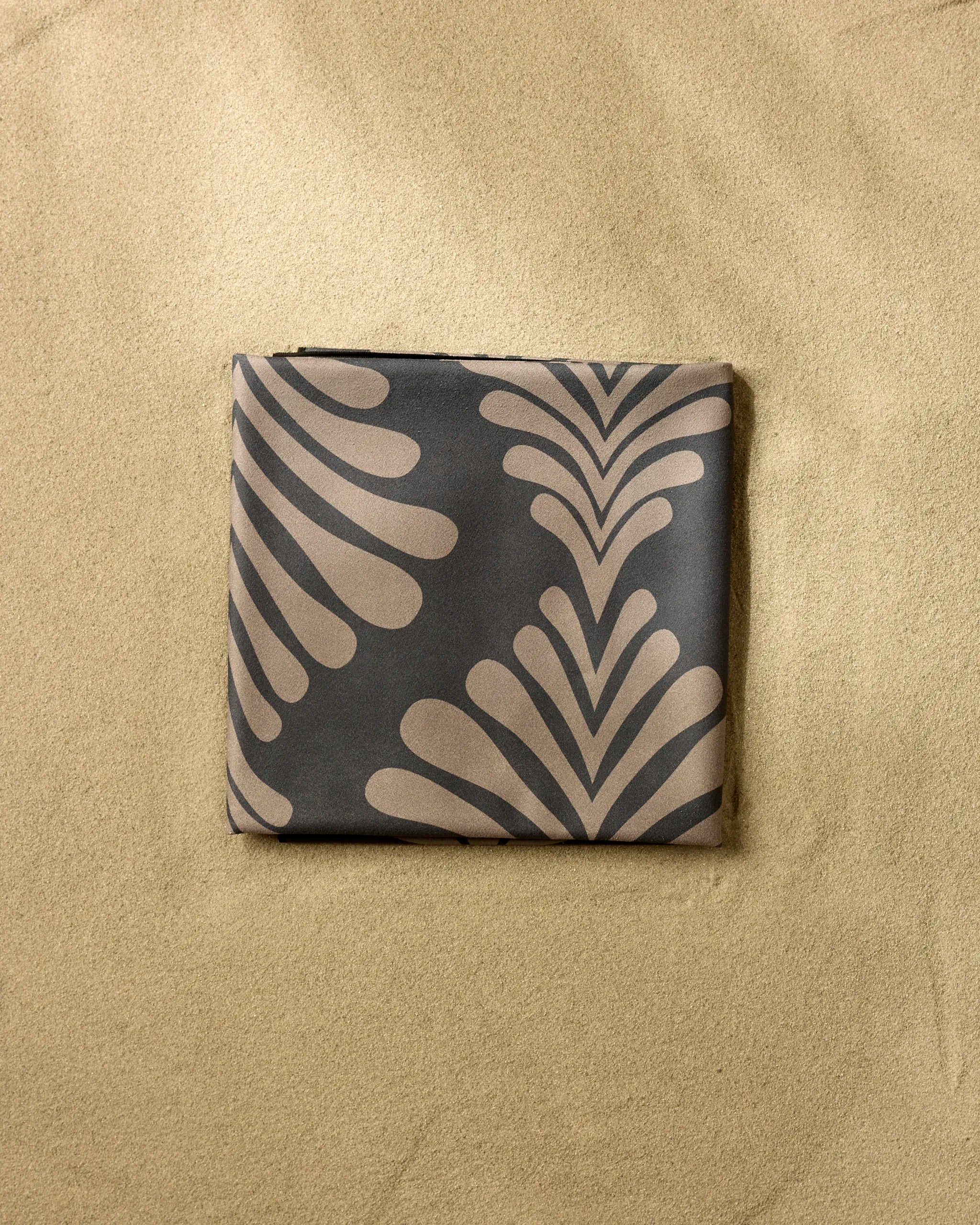 The GEOMETRY Dark Neutrals micro-loop beach blanket, featuring a beige and dark brown abstract leaf design, is neatly folded on the sand.