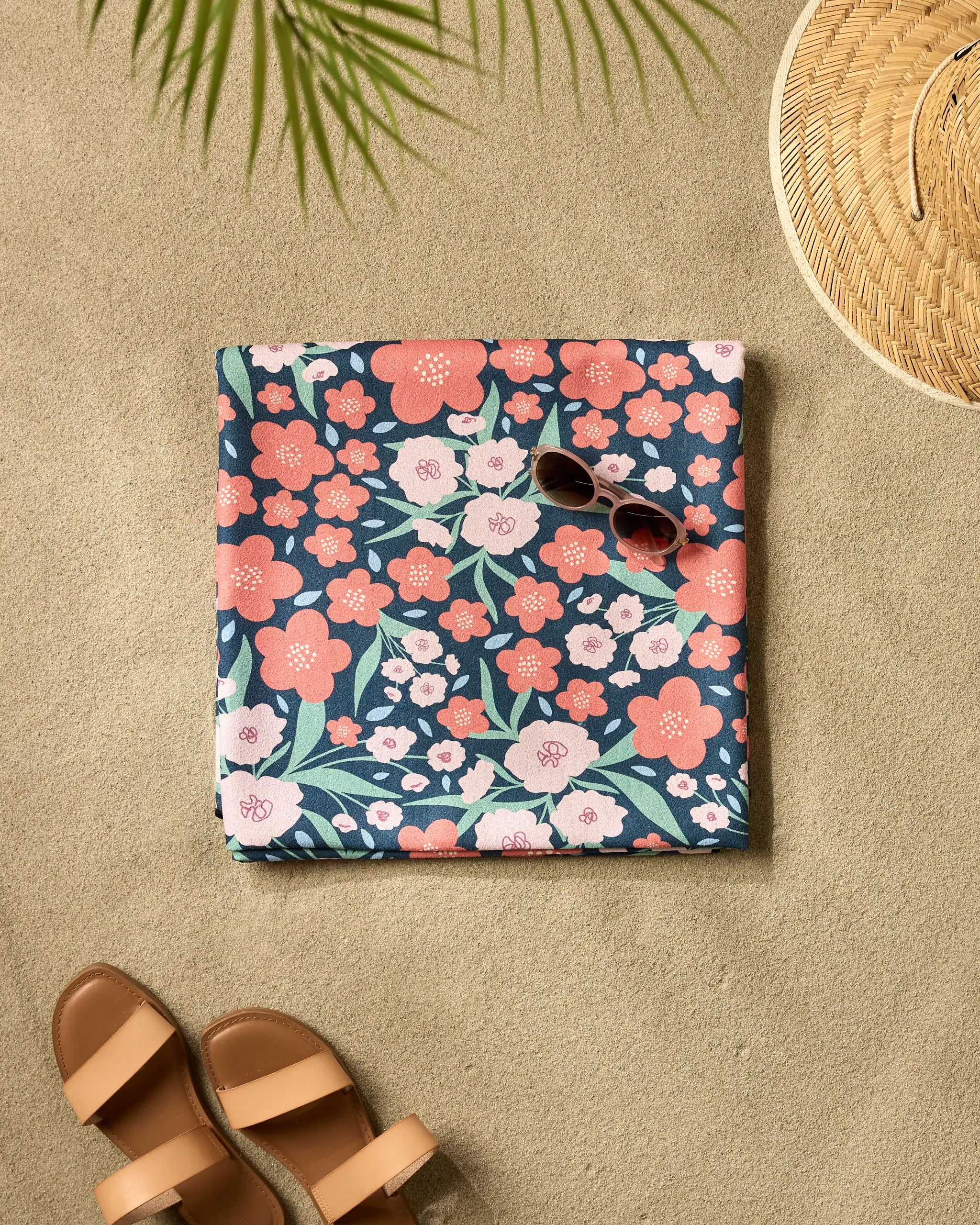 The Daydreamer beach blanket by Charlie Rowan Designs, featuring pink and coral flowers, is spread on the sand with sunglasses on top. Nearby are brown sandals, a woven straw hat, and the shadow of a green palm leaf.