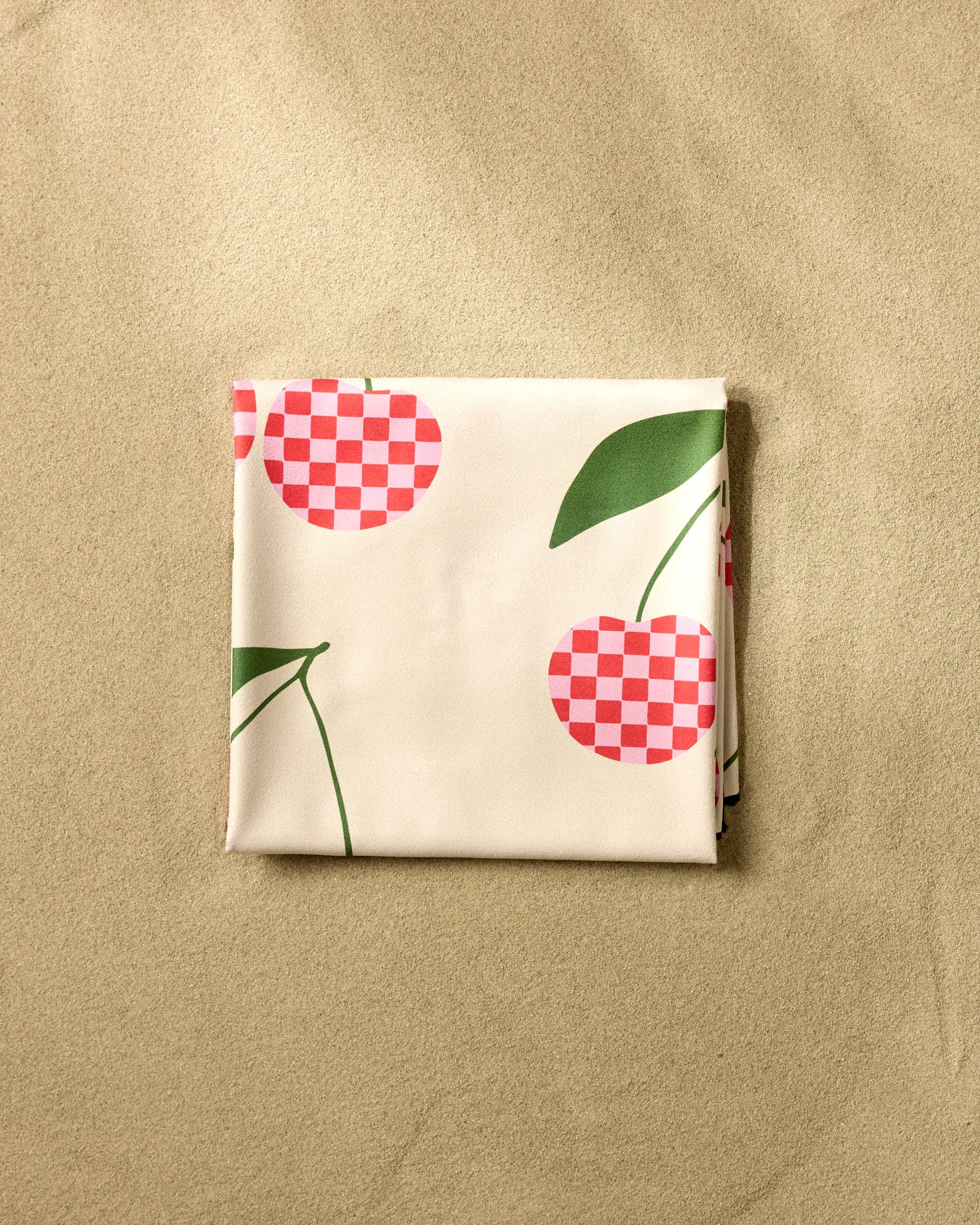 The GEOMETRY Disco Cherry beach blanket features white micro-loop fabric with red checkered cherry patterns and green leaves, shown folded on beige sand.