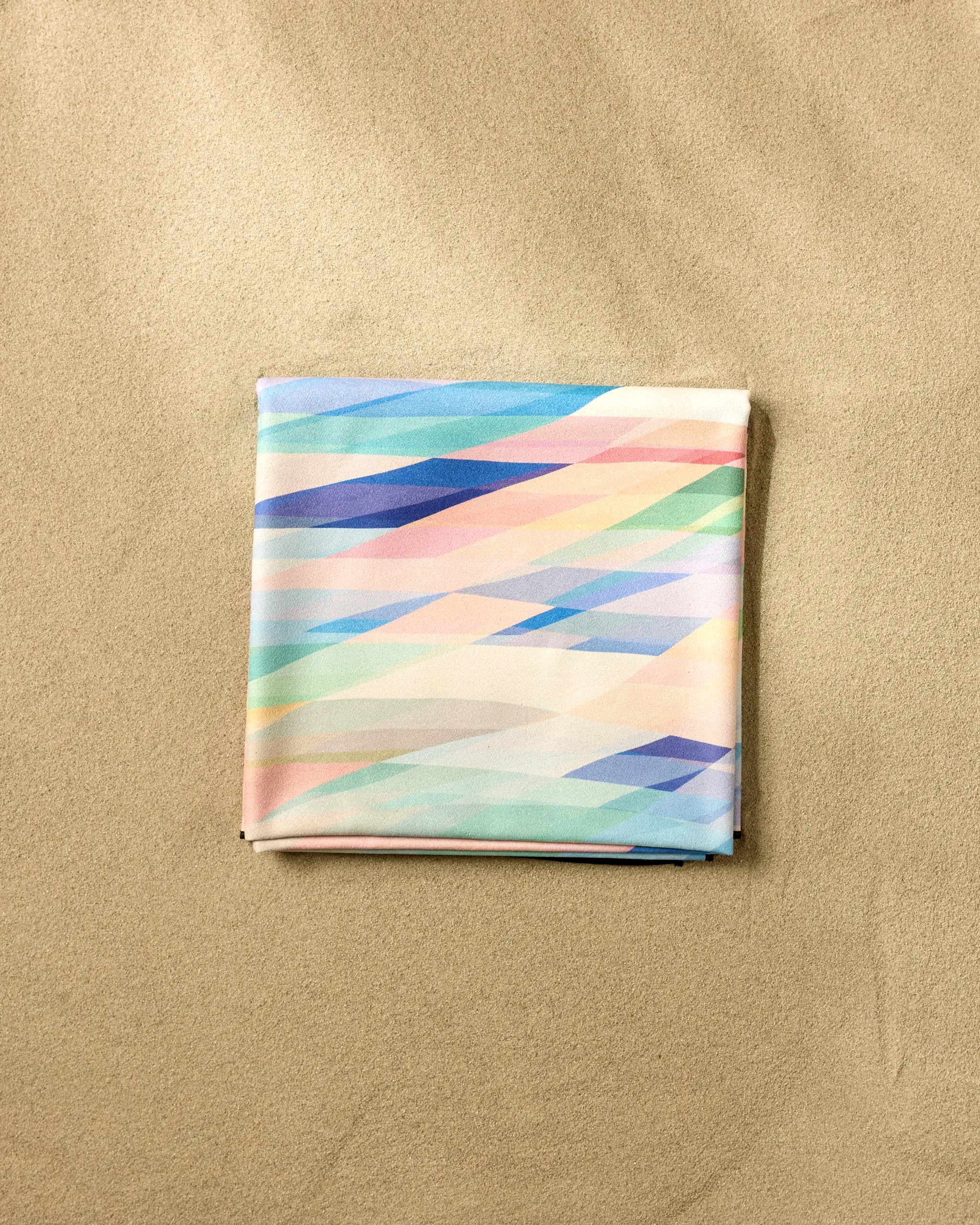 The Eventide square scarf by Rachel Parker features a pastel geometric pattern in blue, pink, green, and cream, neatly folded on a sandy beige surface reminiscent of a soft, quick-drying beach blanket.