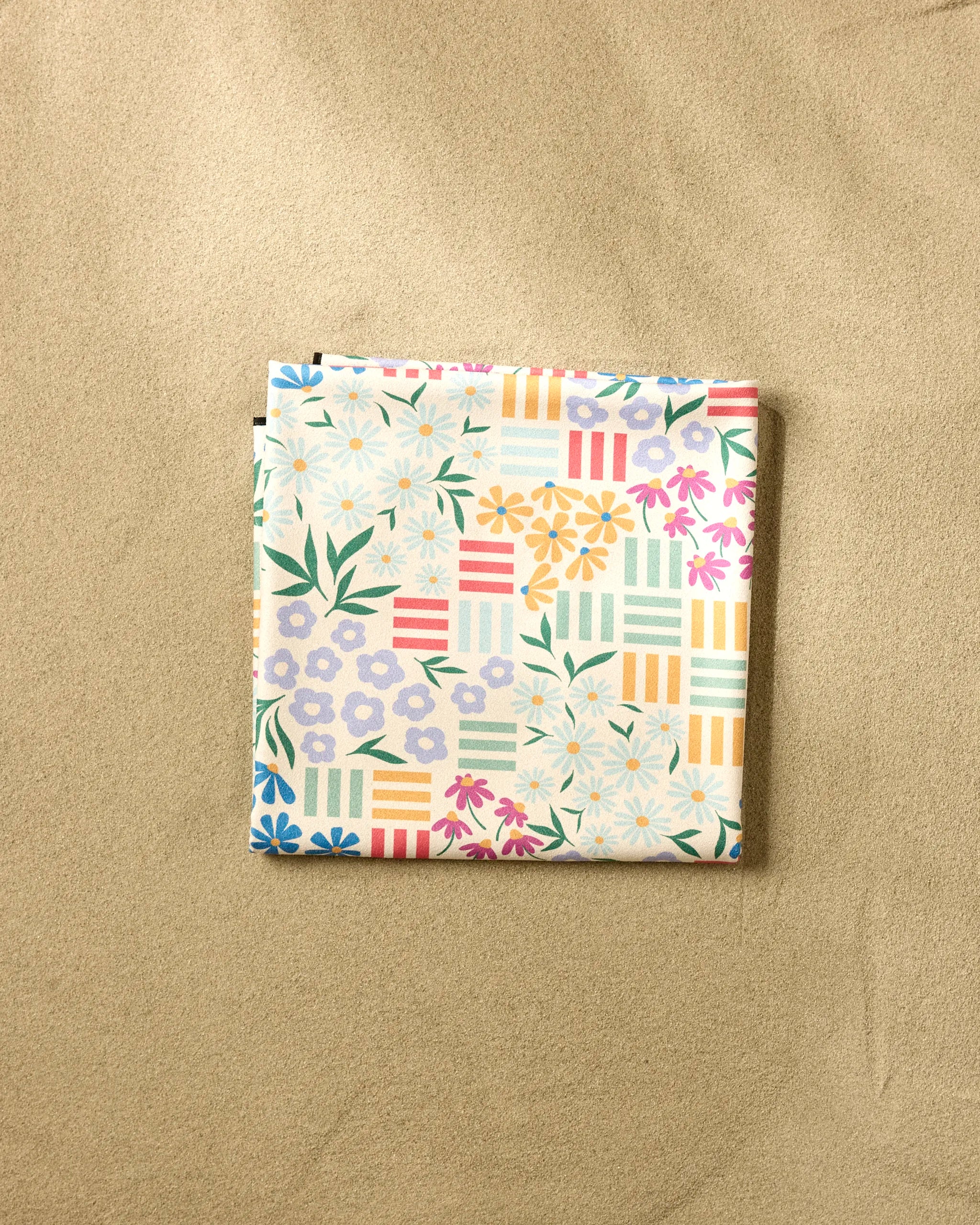 The Floral Garden beach blanket by Charlie Rowan Designs, featuring vibrant floral and geometric prints, is crafted from quick-drying recycled materials and shown neatly folded on sandy beige.