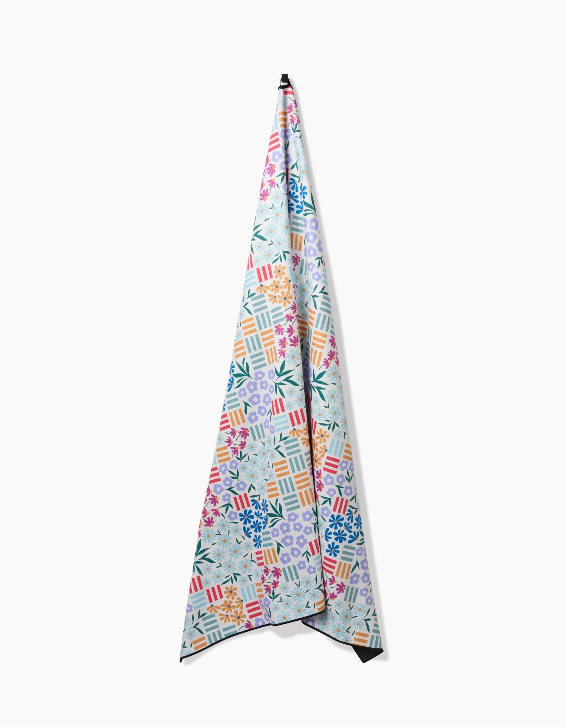 The Floral Garden quick-drying towel by Charlie Rowan Designs, featuring a vibrant geometric and floral pattern, hangs vertically on a white wall with its pointed end facing up.