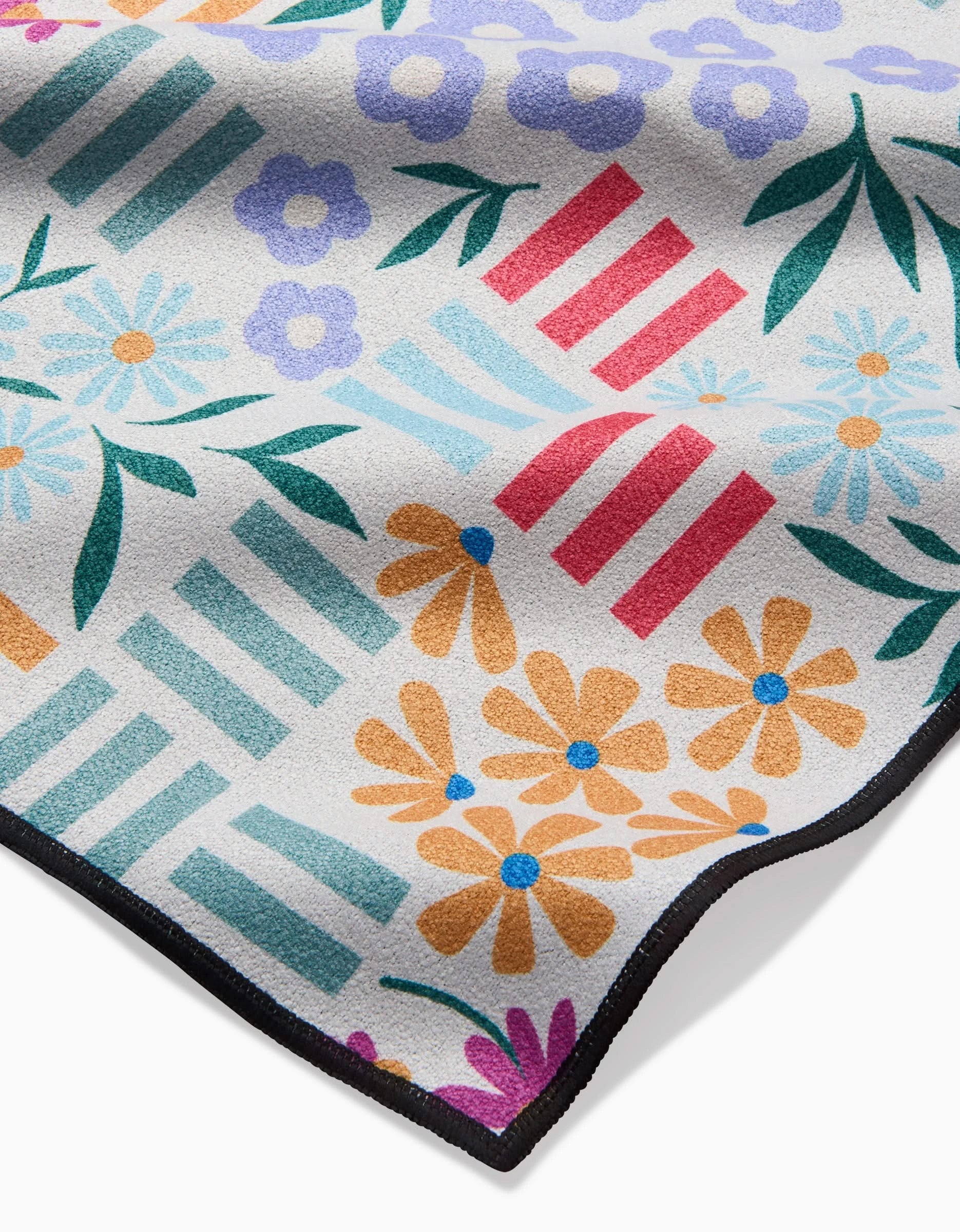 A close-up of Charlie Rowan Designs’ Floral Garden Beach Blanket, made from recycled materials, showcases a vibrant pattern of orange, purple, and blue flowers with green leaves and red and teal stripes on white, finished with a black border.