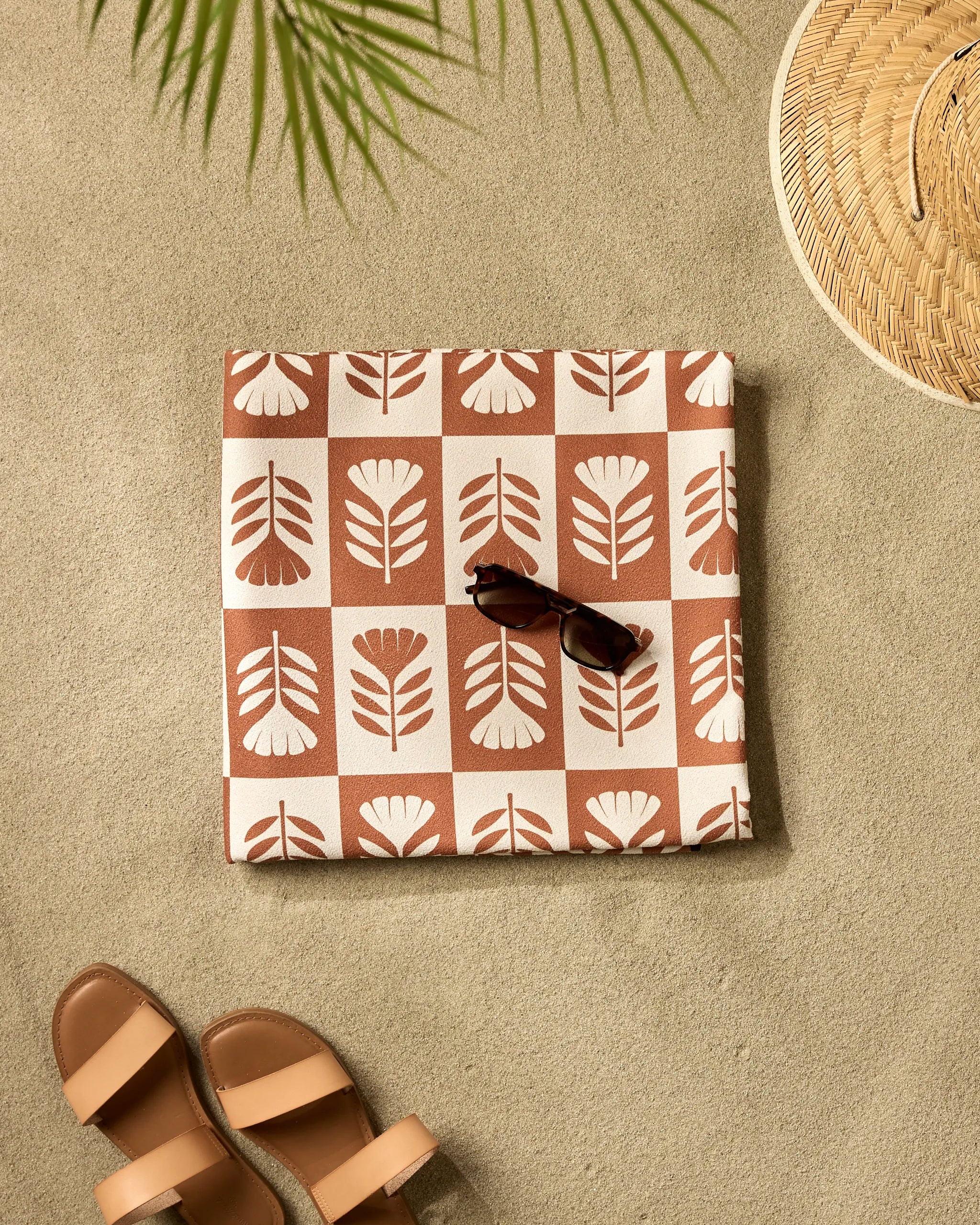 The Flower Patch quick-drying beach blanket from Charlie Rowan Designs, folded with a floral print and sunglasses on top, rests on the sand beside tan sandals, a woven hat, and a palm leaf casting a shadow.