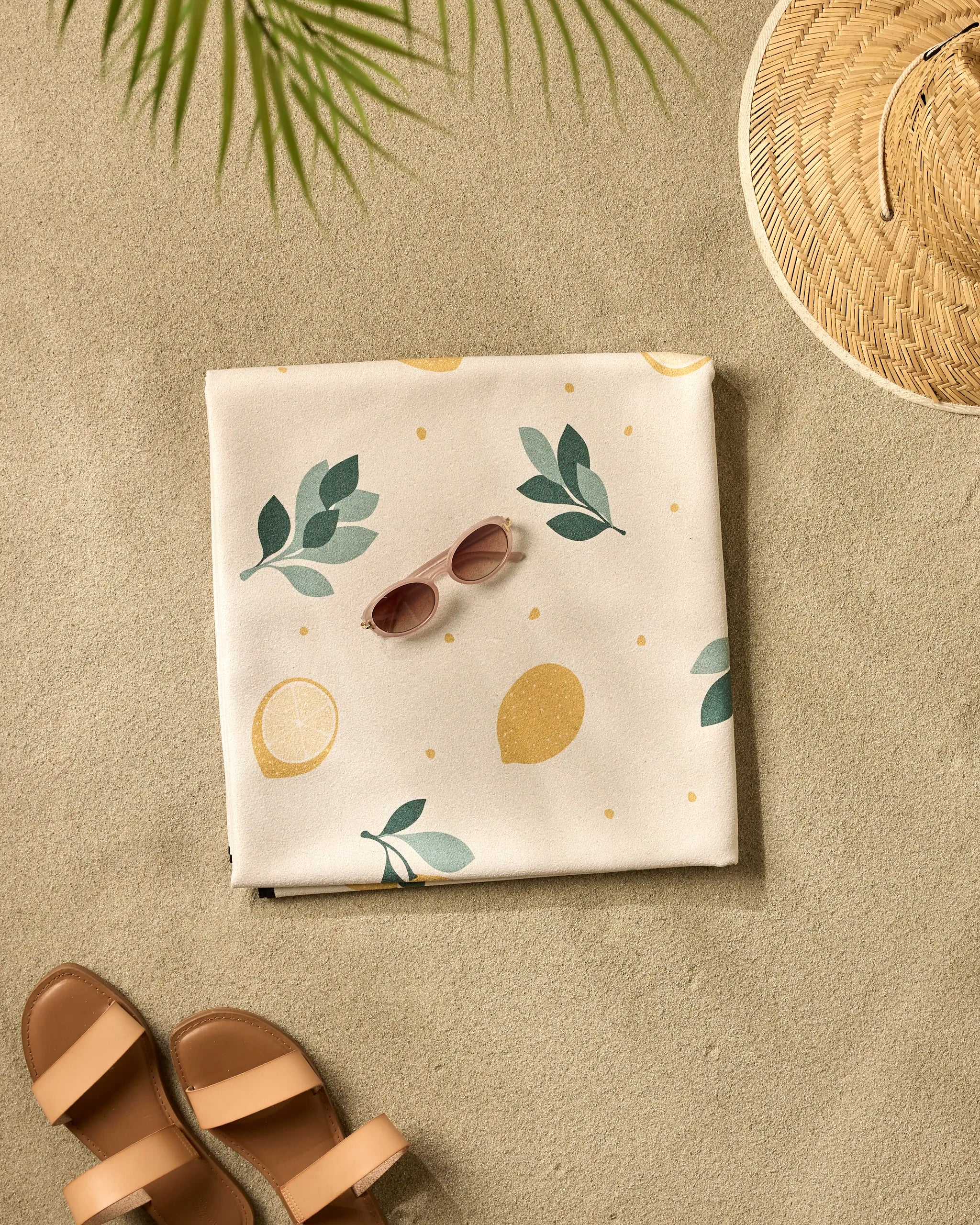 The Sara Mari Freshly Picked micro-loop beach blanket featuring a lemon and leaf pattern is spread on the sand with pink sunglasses, a straw hat, and brown sandals beneath the shadow of a palm leaf.