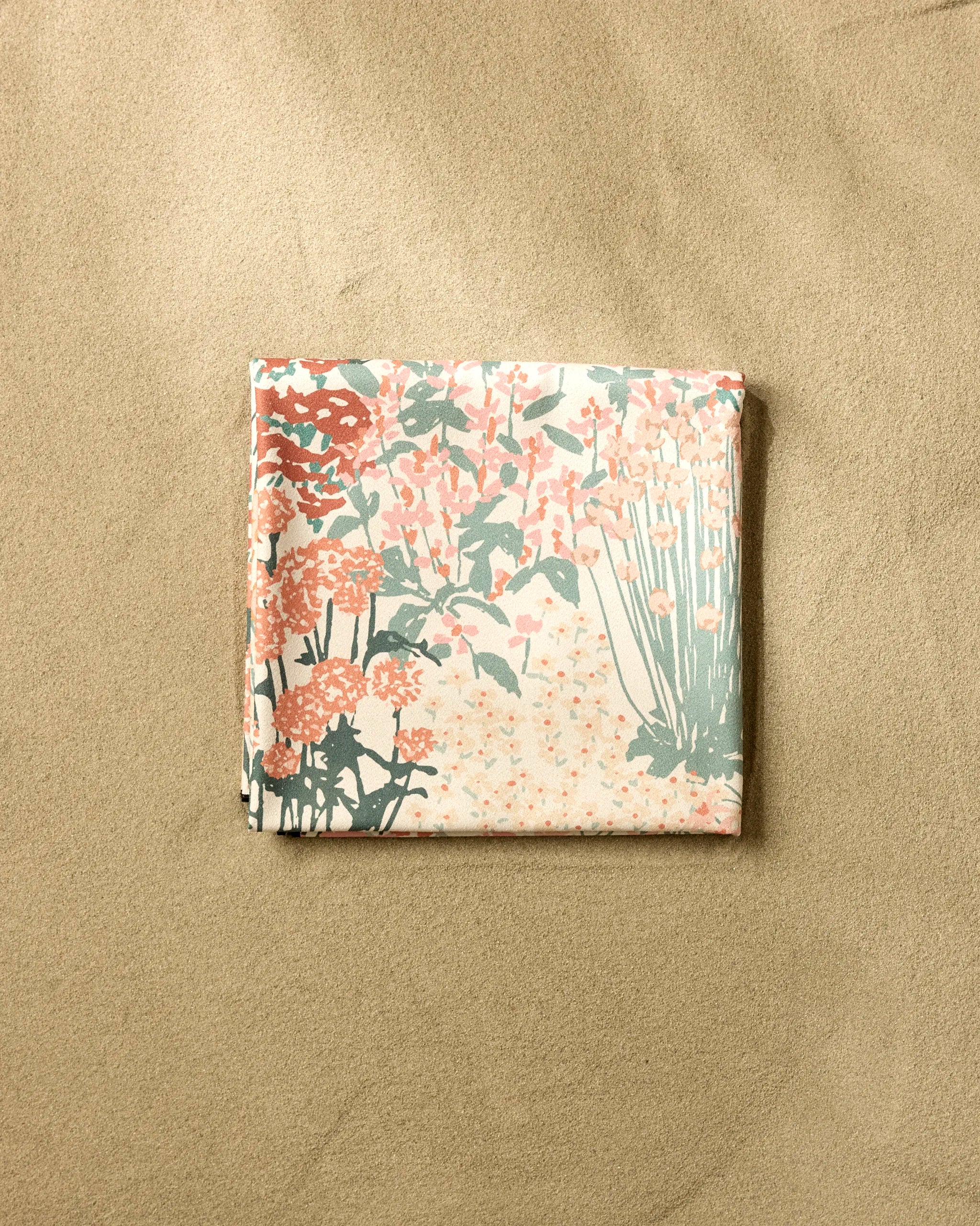 The Garden Bloom beach blanket by Hufton Studio, featuring a pink, green, and cream floral pattern and sand-repelling fabric, is folded on a textured tan surface.