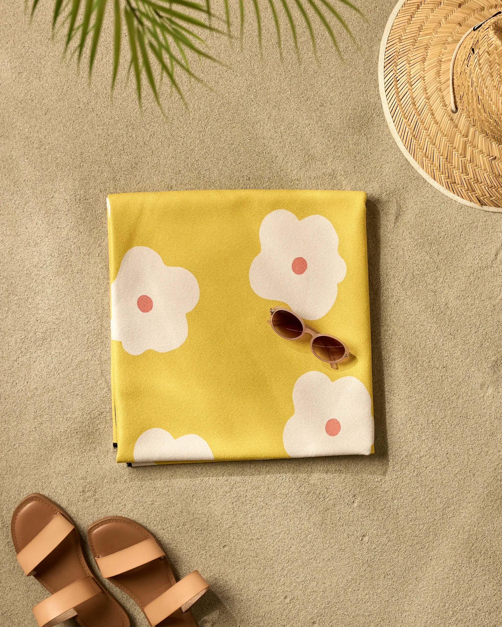 The Sara Mari Garden Crush yellow beach blanket, featuring white and pink floral designs and made from quick-drying recycled materials, lies on the sand beside brown sandals, round sunglasses, a straw hat, and a shadowy palm leaf.