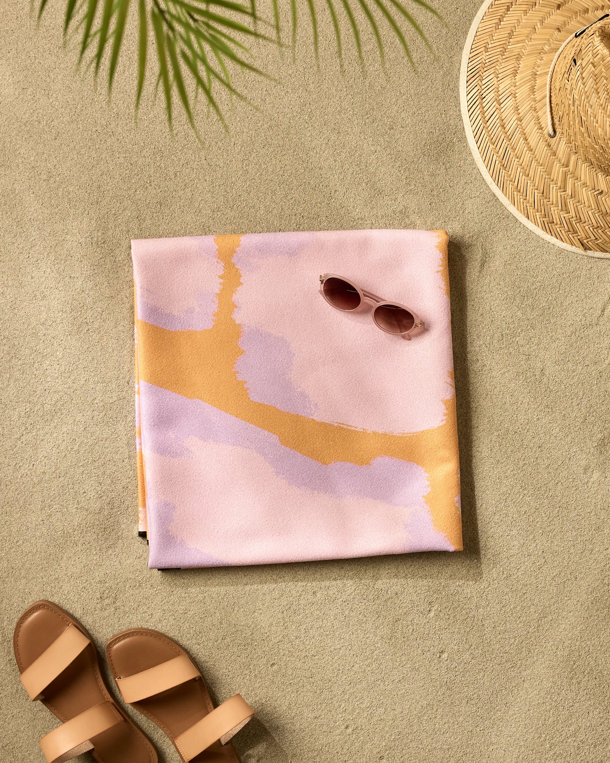 A folded Gentle Giraffe quick-drying beach blanket by Elisabeth Wing in pastel pink and orange lies on sandy ground, surrounded by sunglasses, sandals, a woven sunhat, and a palm leaf shadow above.