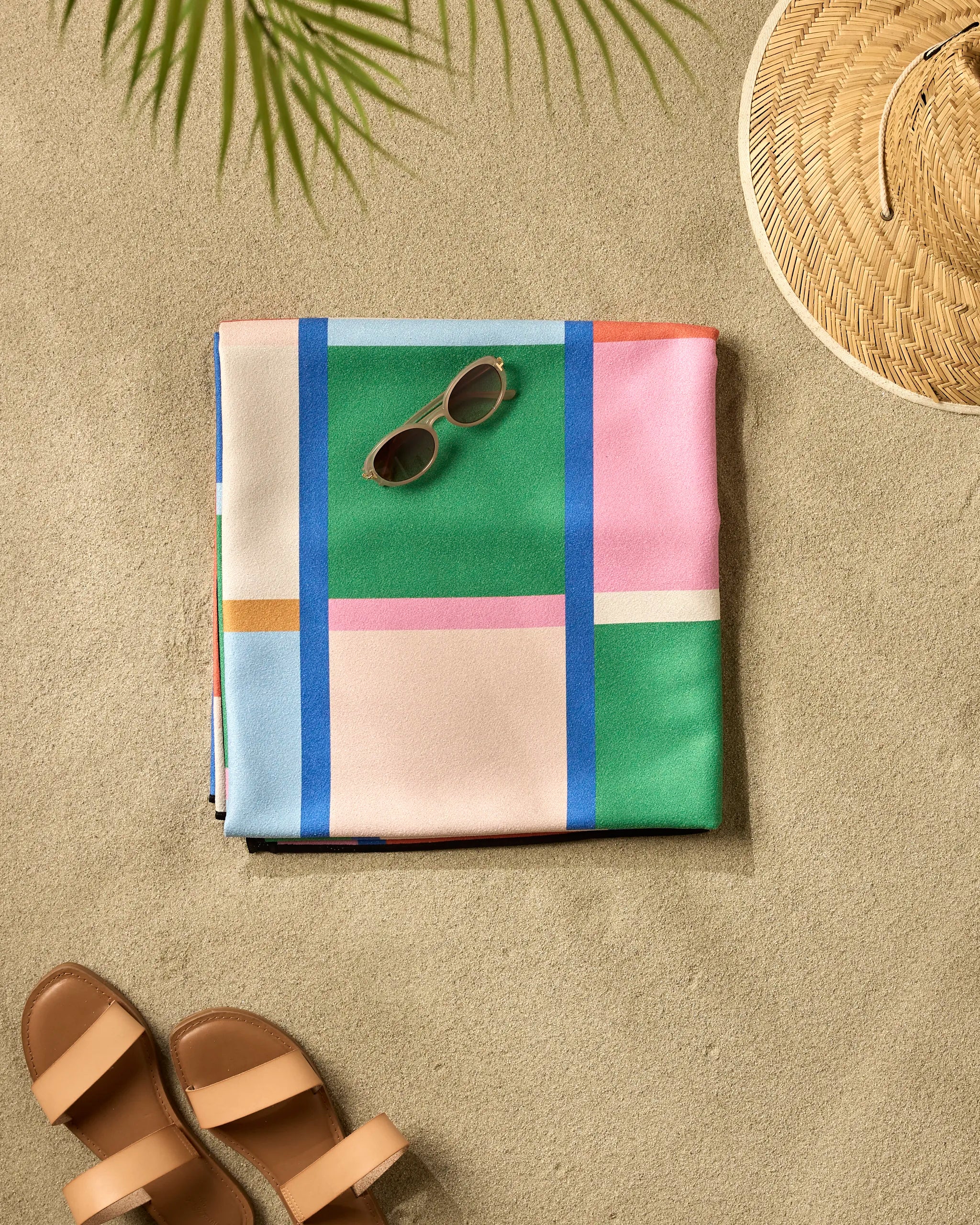 The Jennifer Bouron Grid recycled beach blanket, with a colorful checkered design, is folded on the sand alongside sunglasses, a straw hat, and brown sandals beneath the shadow of a palm.