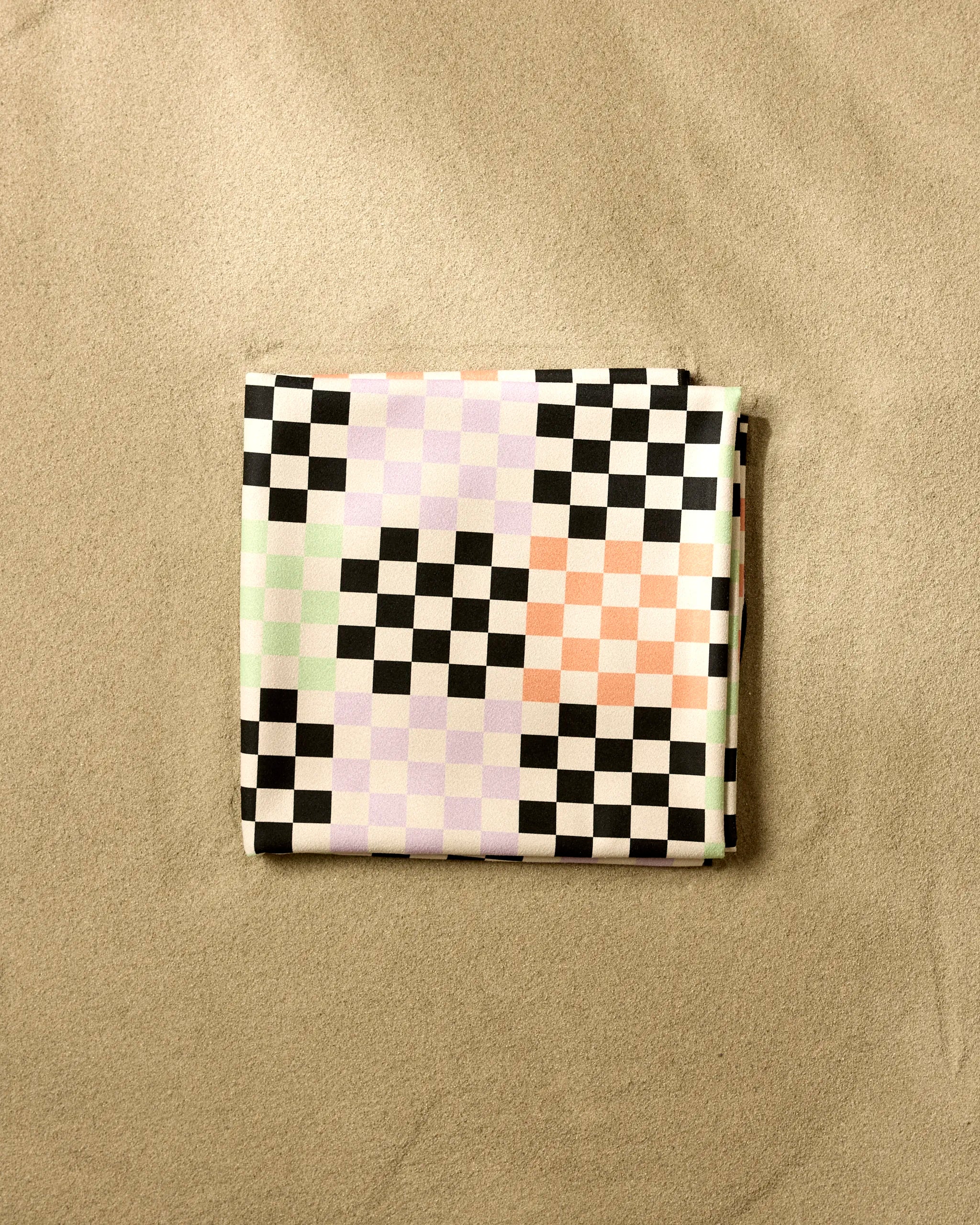 The GEOMETRY Hit Block is a quick-drying beach blanket featuring a checkerboard pattern in black, white, pale pink, and light green squares, shown resting on sand.