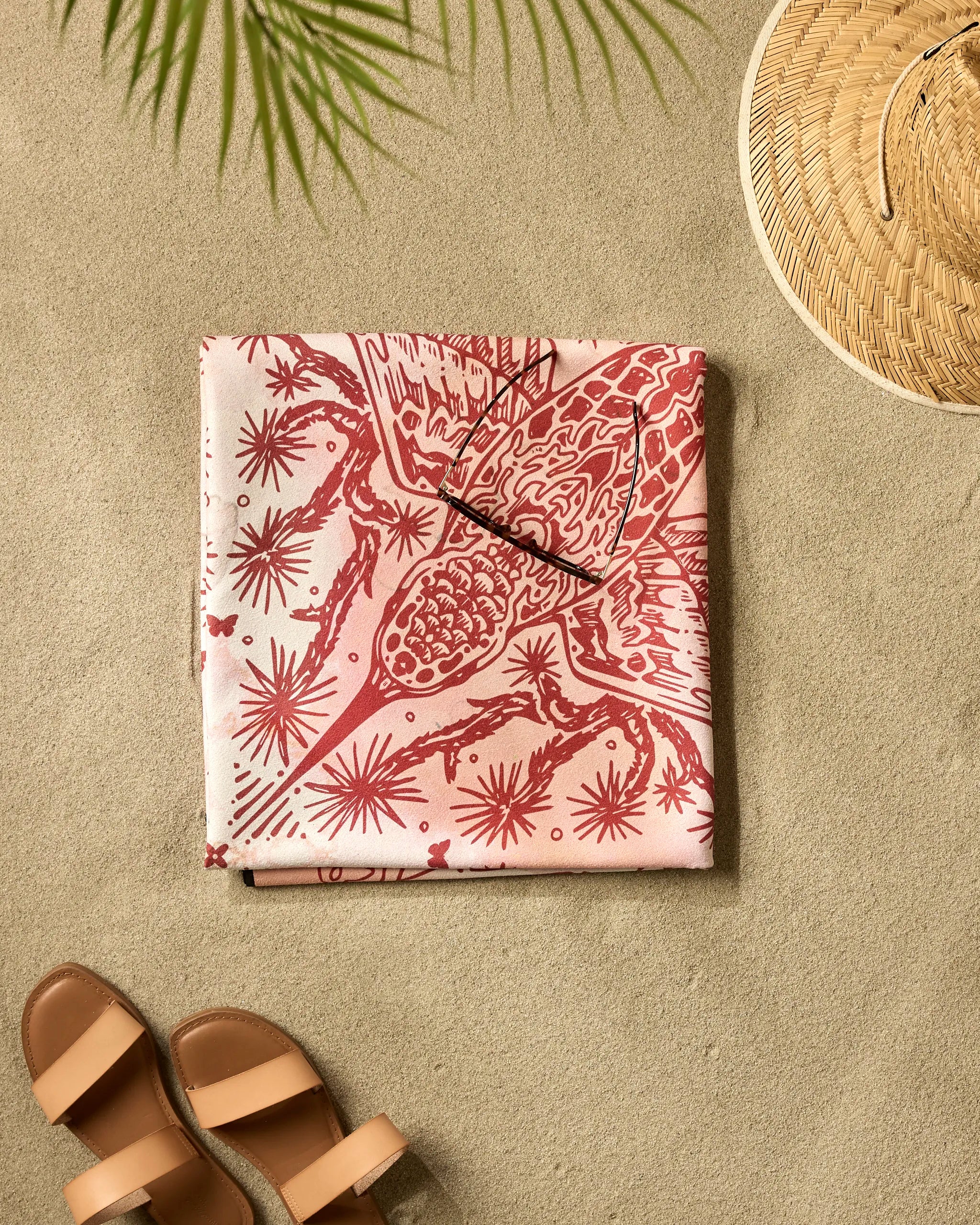 The Skye Walker Hummingbird Beach Blanket, folded in pink and red micro-loop fabric, rests on the sand next to brown sandals, a straw hat, and palm leaves casting their shadows.