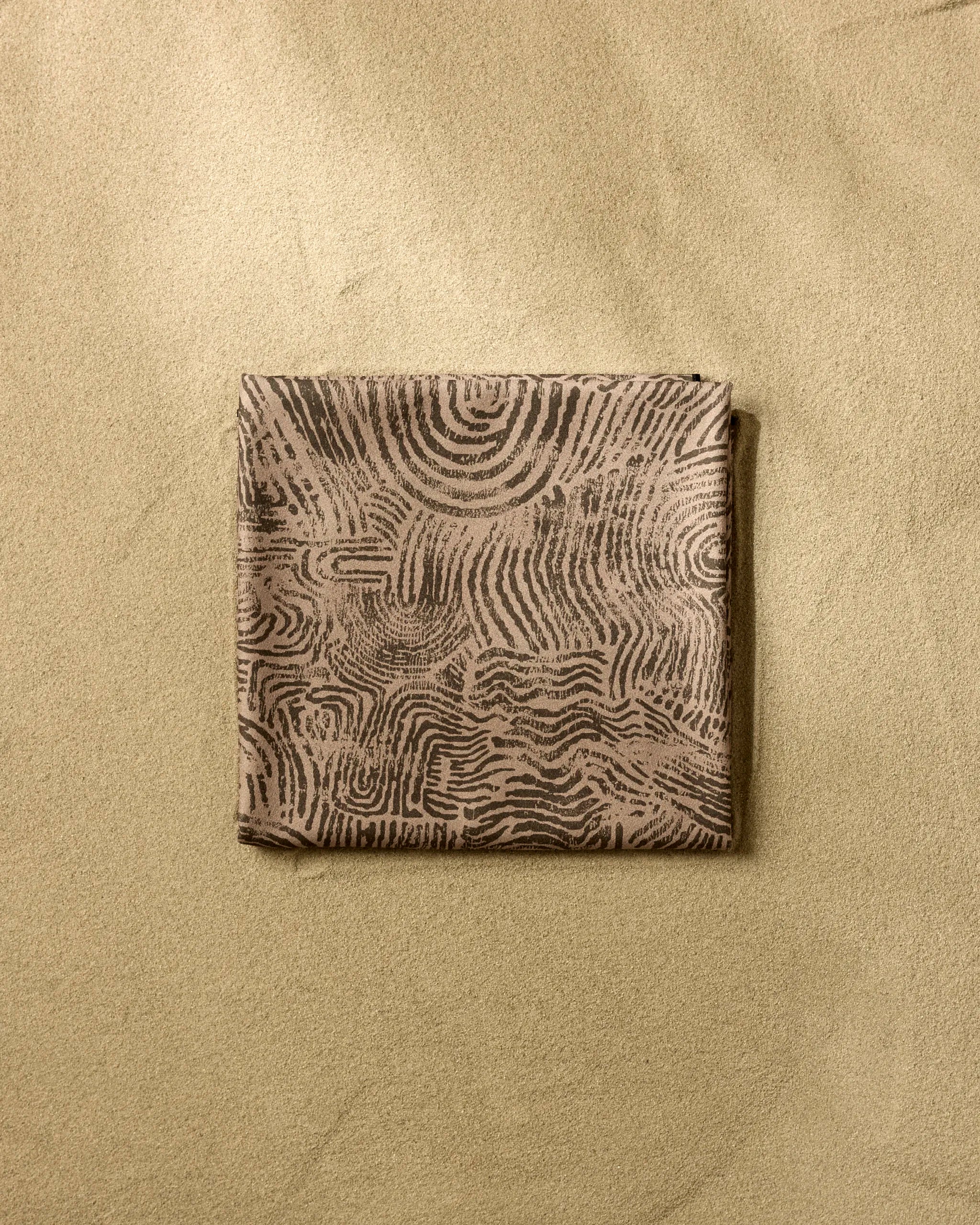 The Imprint beach blanket by Studio Jeannot is square, quick-drying, and features an abstract wavy dark brown pattern on a light brown background, shown here on a sand-colored textured surface.