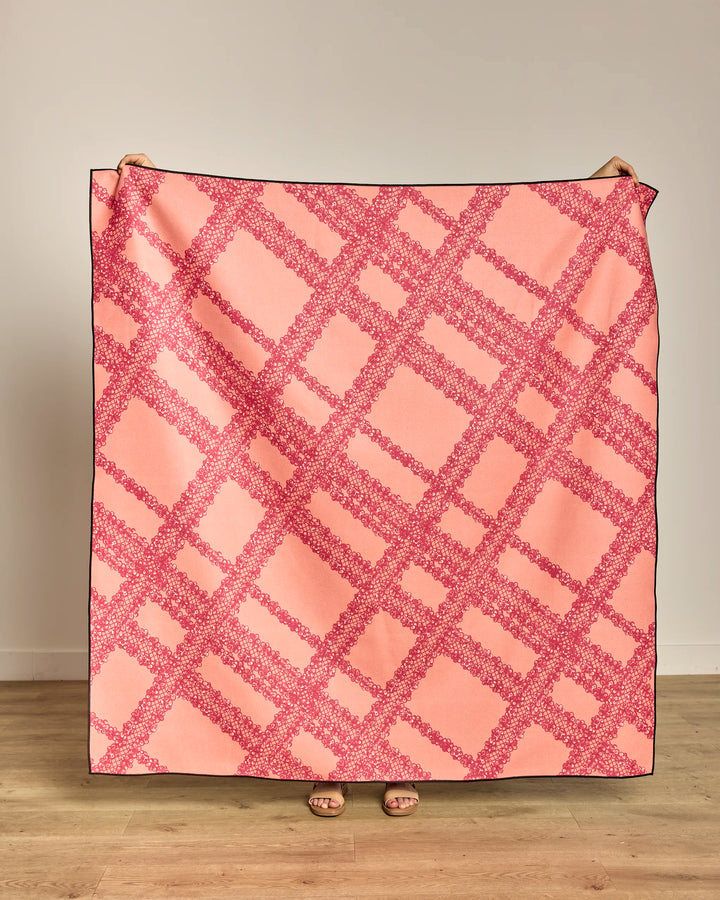 The Geometry Lacy Plaid beach blanket, in large pink and red plaid, is held upright to cover most of a person—only hands at the top and sandal-clad feet below show—against a plain wall with a wooden floor.