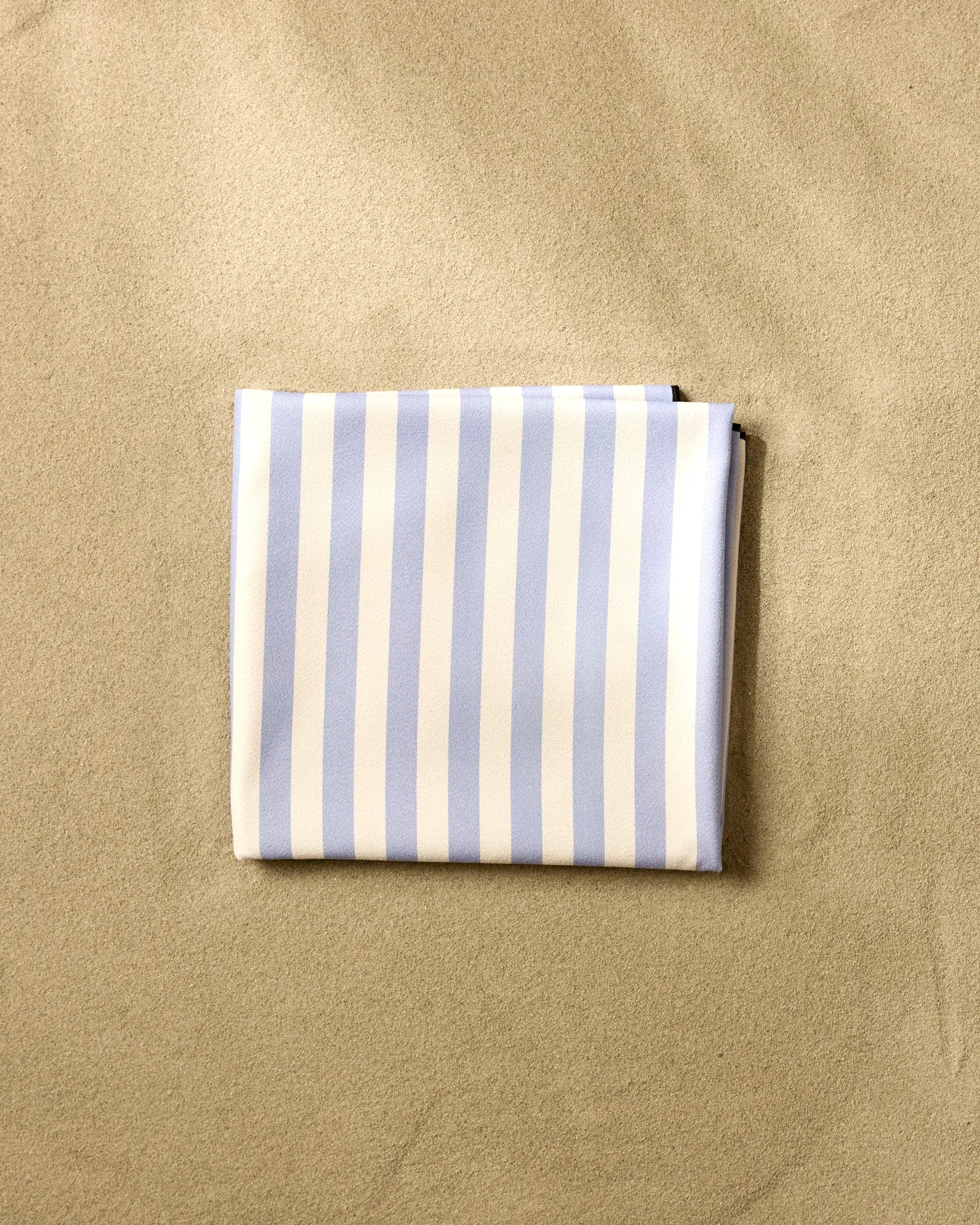 The GEOMETRY "Light Blue Stripes" micro-loop beach blanket, neatly folded, sits on smooth, pale sand.