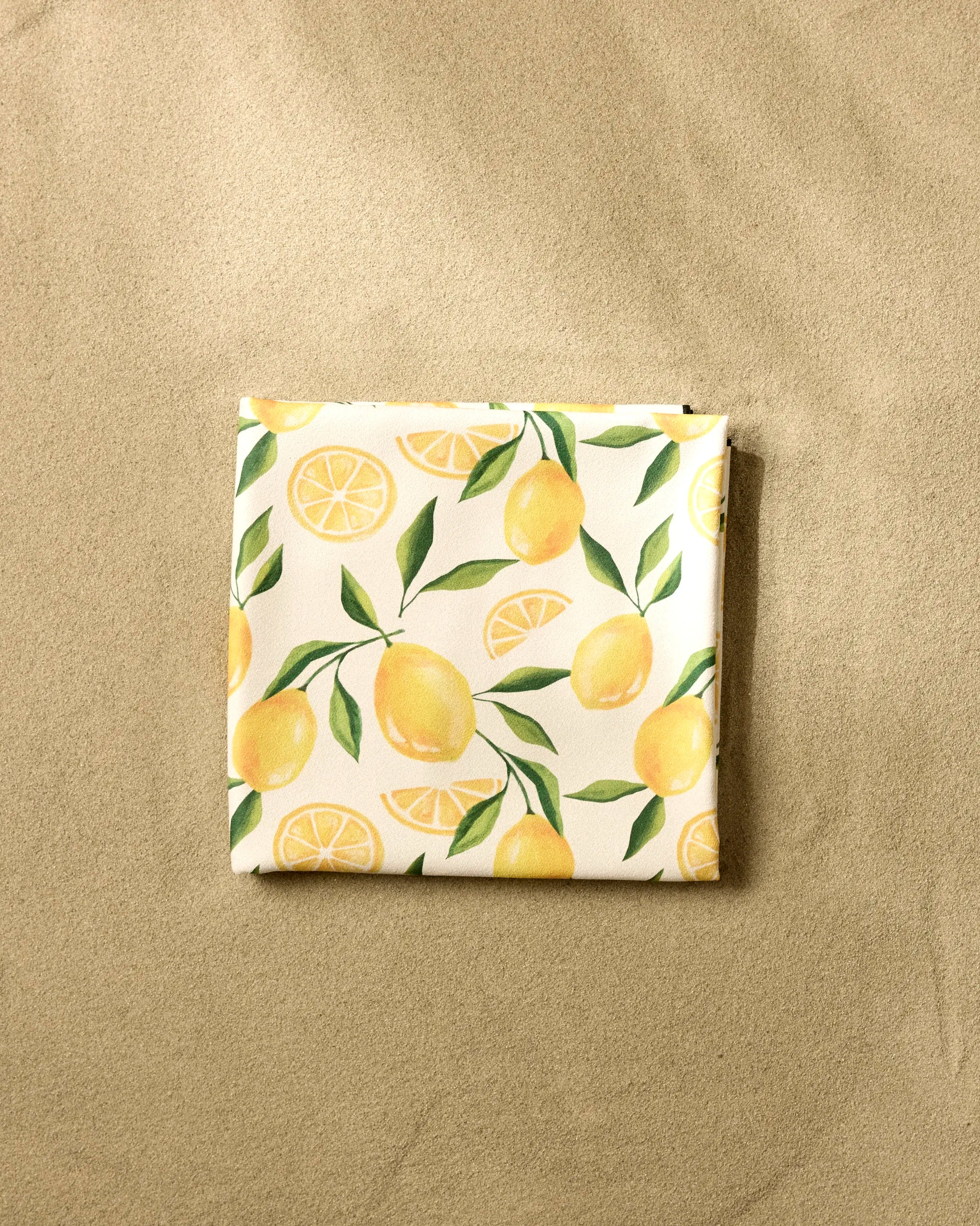 The GEOMETRY Limoncello beach blanket, crafted from quick-drying recycled materials, features a lemon and leaf pattern and is shown folded on smooth sand.