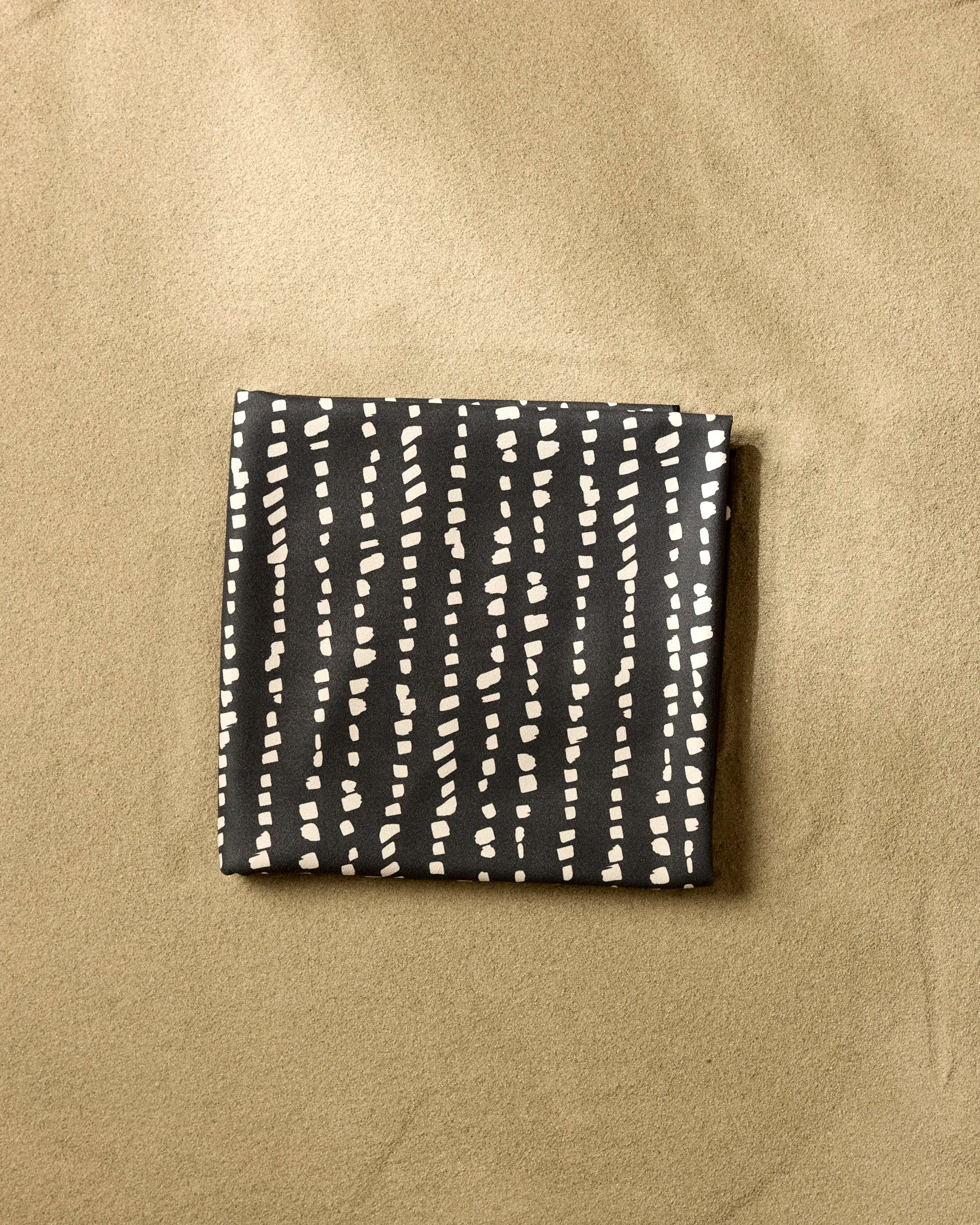 The GEOMETRY Little Prints is a folded, dark beach blanket crafted from recycled materials with irregular white dotted lines, shown resting on sand.