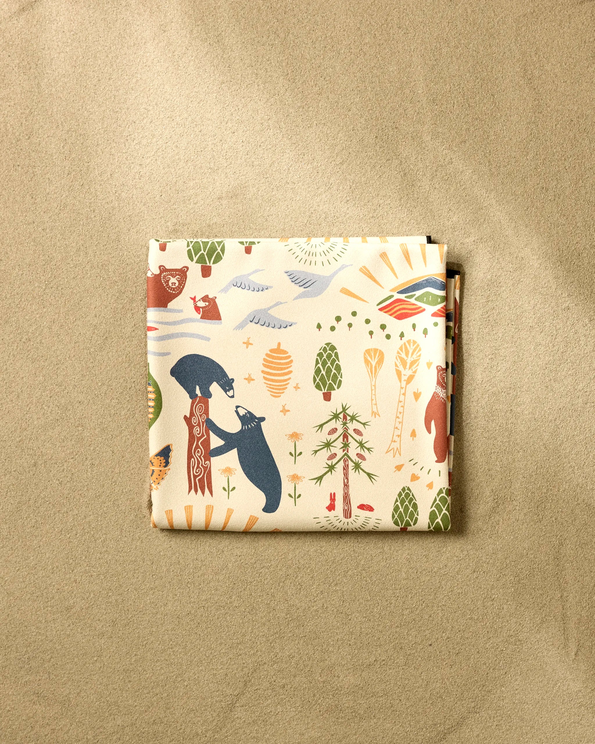 The "Mama Bear" folded beach blanket by Elisabeth Wing, crafted from recycled materials and featuring bears, trees, pinecones, and sun motifs in earthy tones, rests on a sandy beige background.