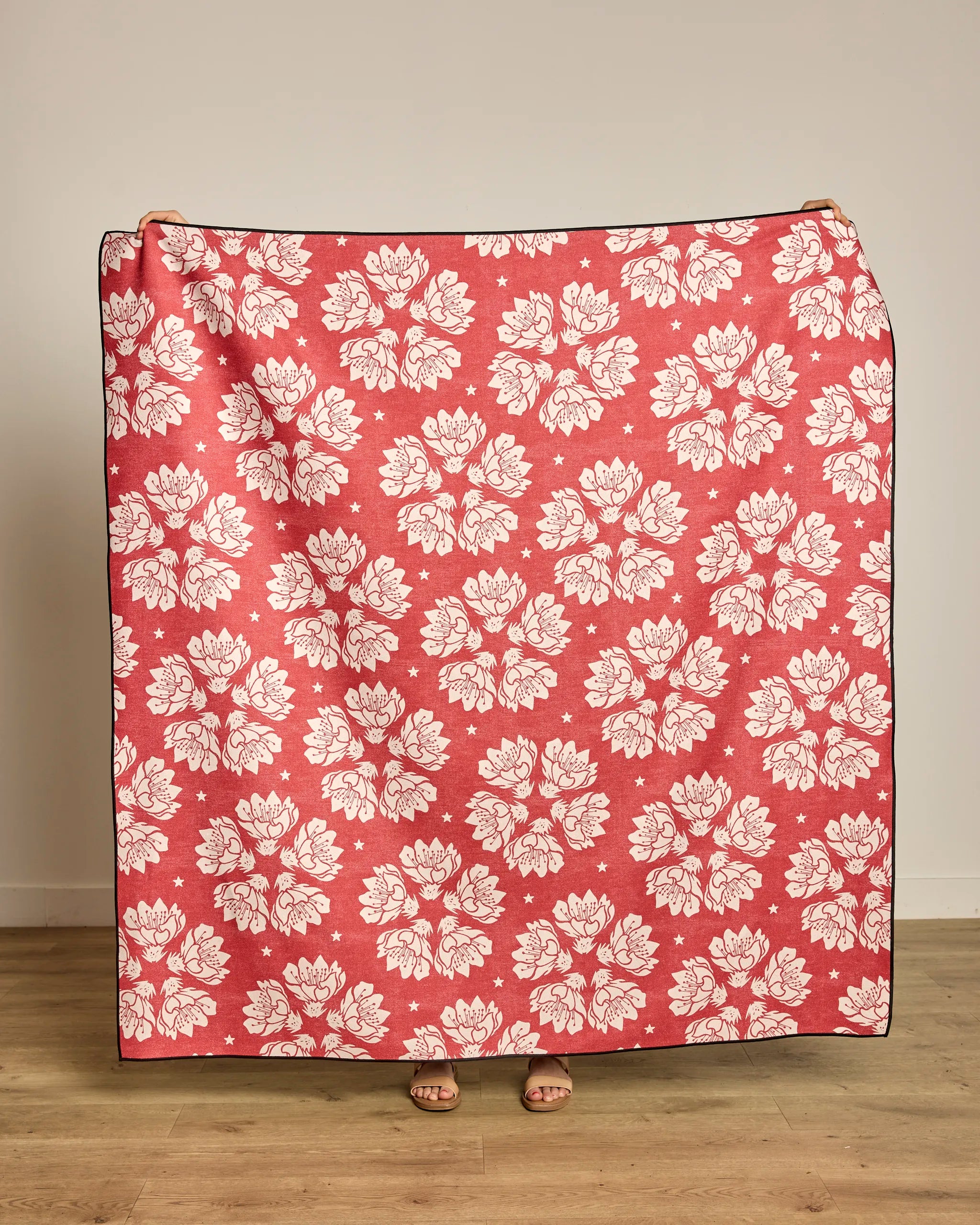A person holds up the Maverick Floral beach blanket by Geometry, featuring a large pink and white floral pattern. The quick-drying, recycled material stands out against the beige wall and wooden floor behind them.