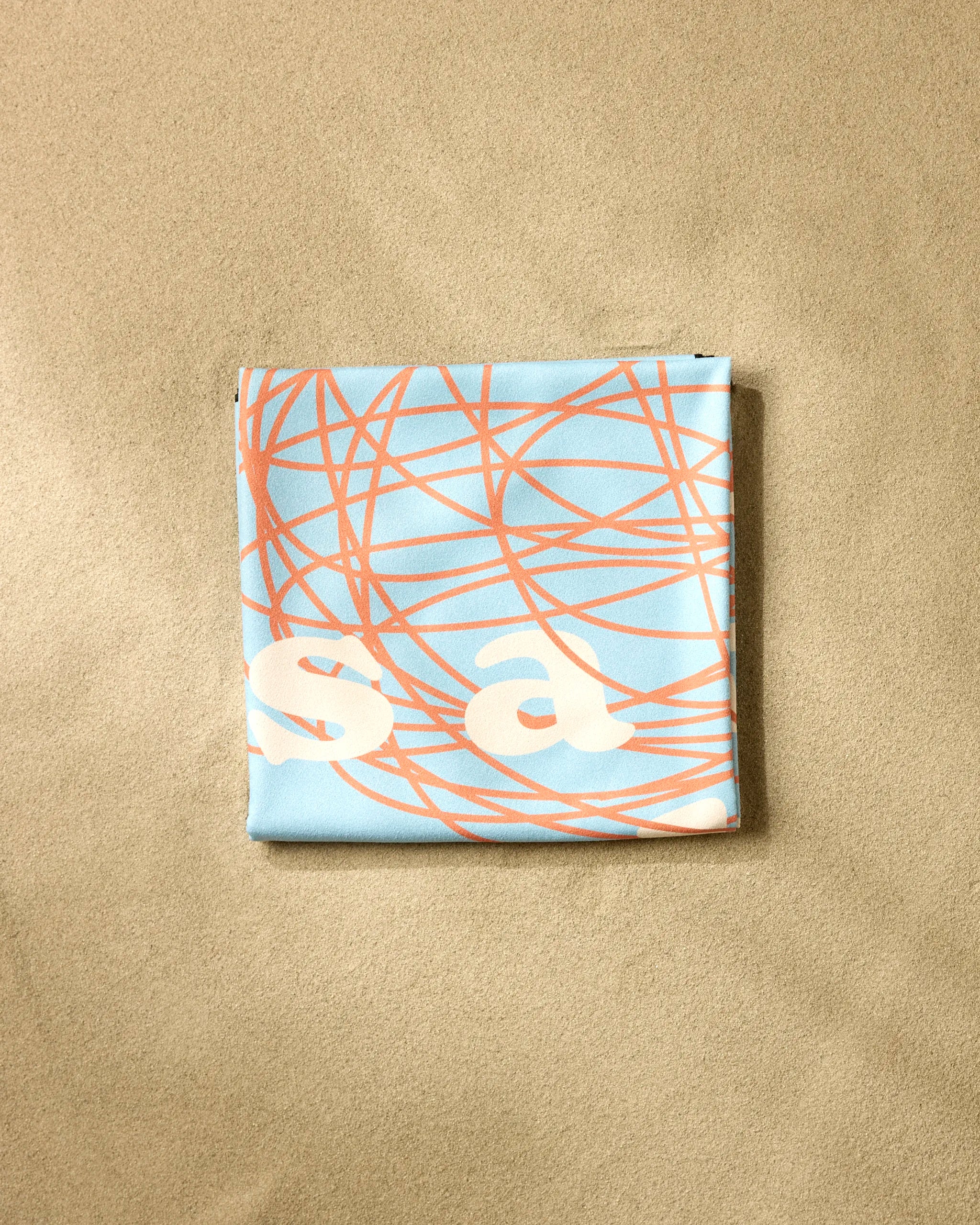 A folded Messy Life beach blanket by Quotes by Christie, featuring blue and beige with abstract orange lines and partial white letters, rests on a light tan textured surface.