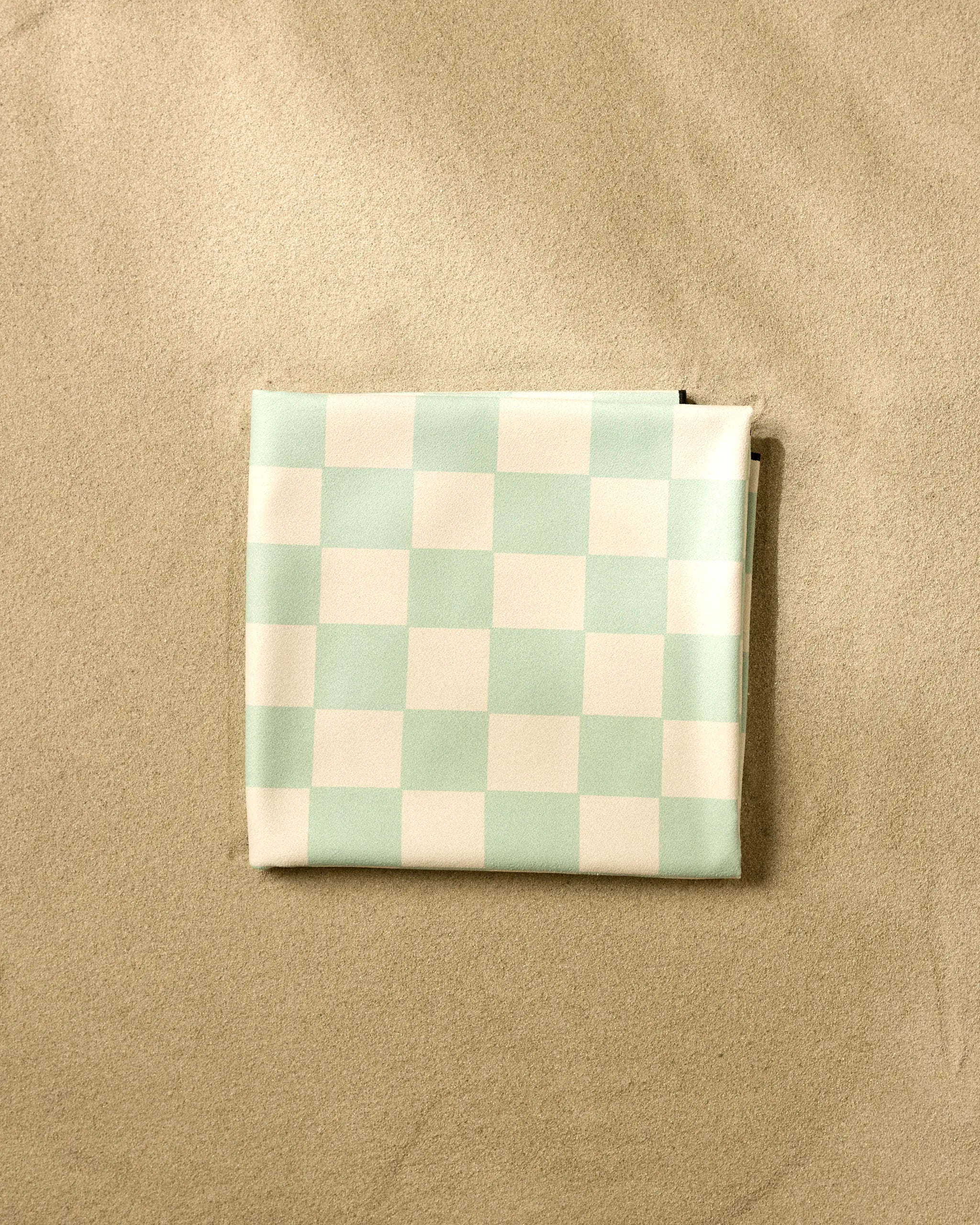 A Mint Checker pouch by GEOMETRY, featuring light blue and white checks with a zipper, is placed on light tan sand and viewed from above.