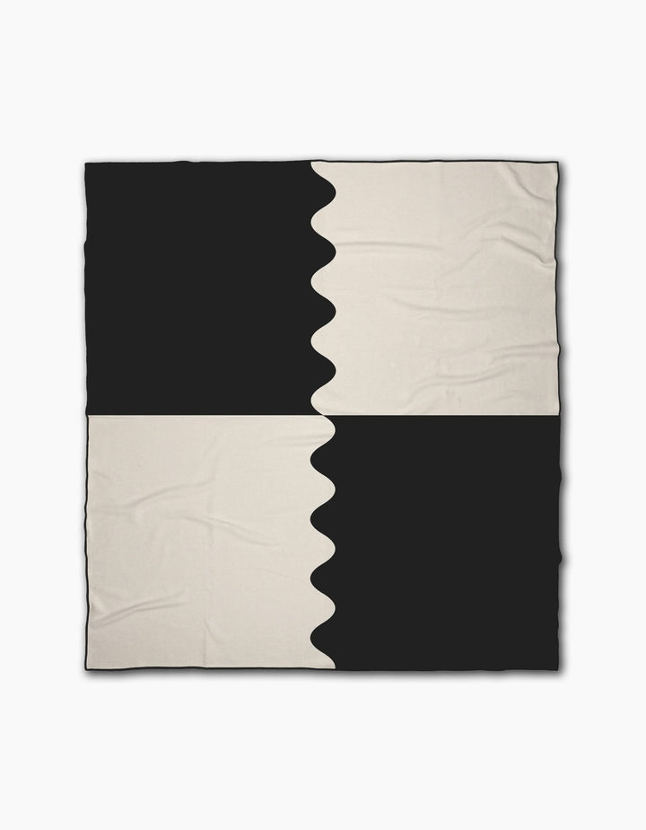 The Geometry Monochrome Waves beach blanket features a black and off-white checkered pattern with smooth and wavy color divisions for an abstract look. Made from quick-drying recycled materials, it has a slightly wrinkled texture.