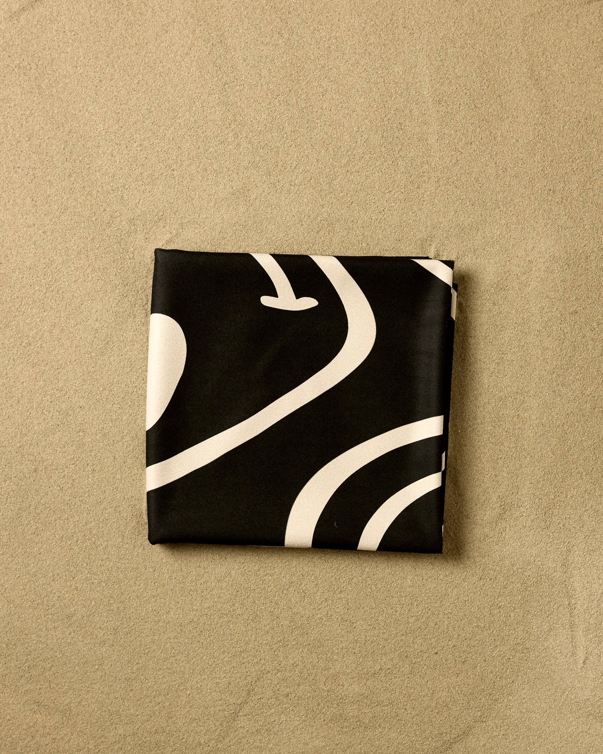 The Mood Swing Dark beach blanket by GEOMETRY, featuring quick-drying black fabric with white abstract lines, is neatly folded into a square on a textured beige surface.
