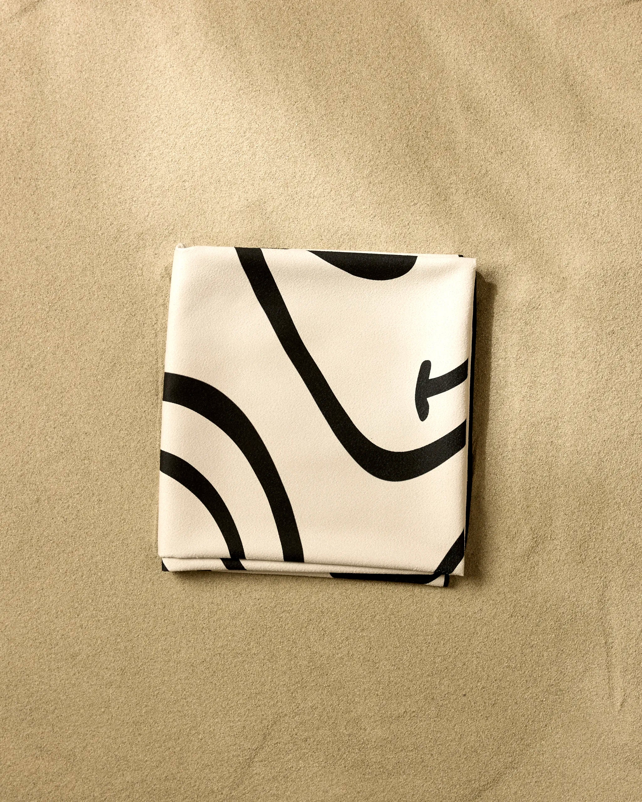 The GEOMETRY Mood Swing Light is a folded white beach blanket with bold black abstract lines, crafted from quick-drying, recycled materials, and shown lying on smooth sand.
