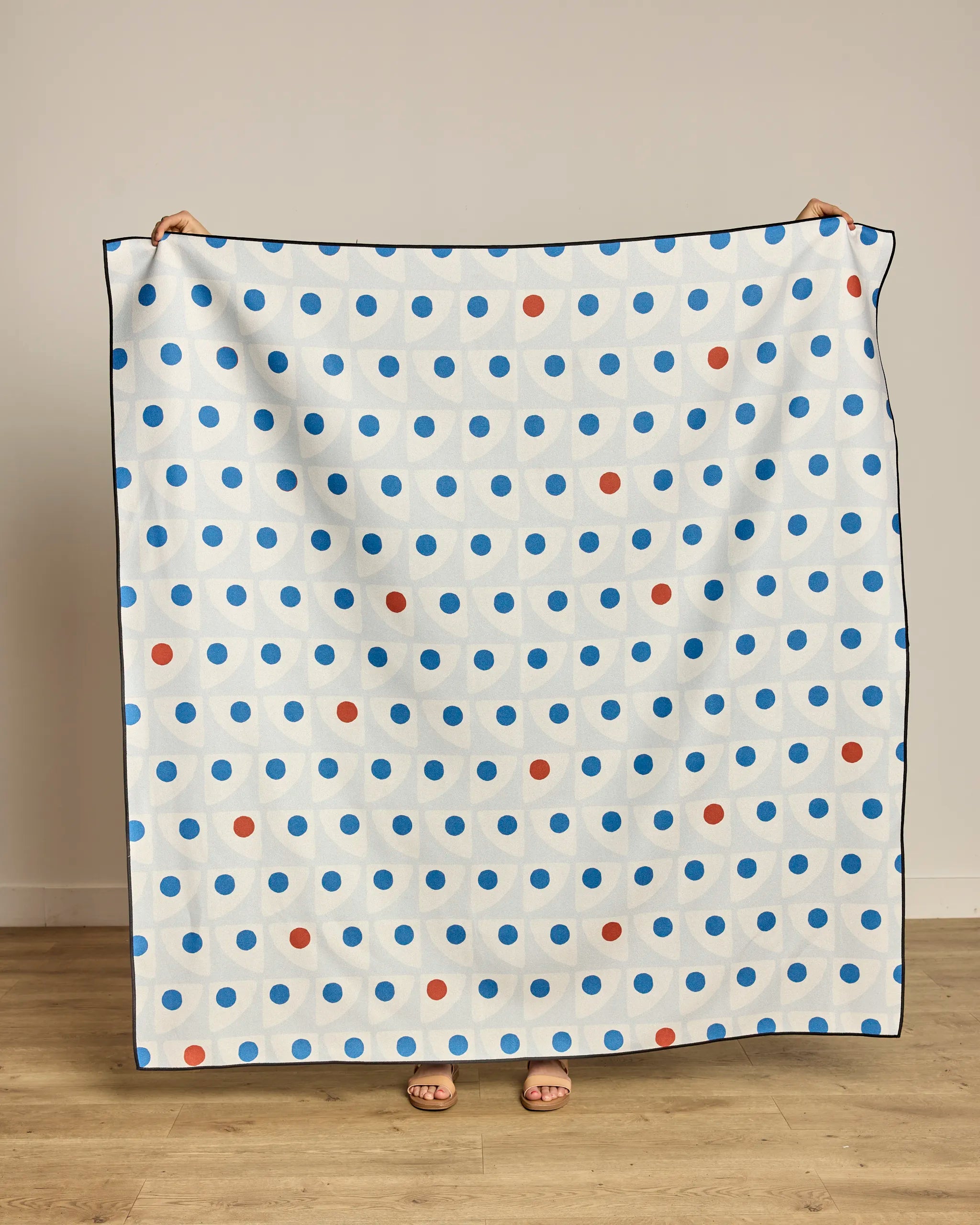 A person stands against a plain wall holding up a large white Geometry Nautical Eye Beach Blanket; only their hands and feet are visible.