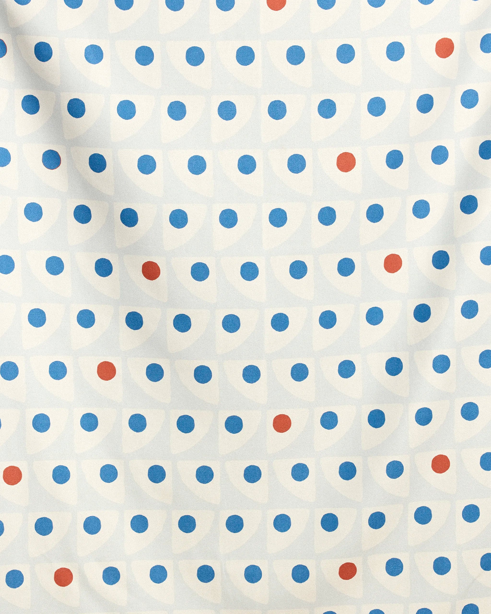The Geometry Nautical Eye Beach Blanket showcases a repeating geometric blue and red dot pattern on white, with subtle folds and shadows adding texture and depth.