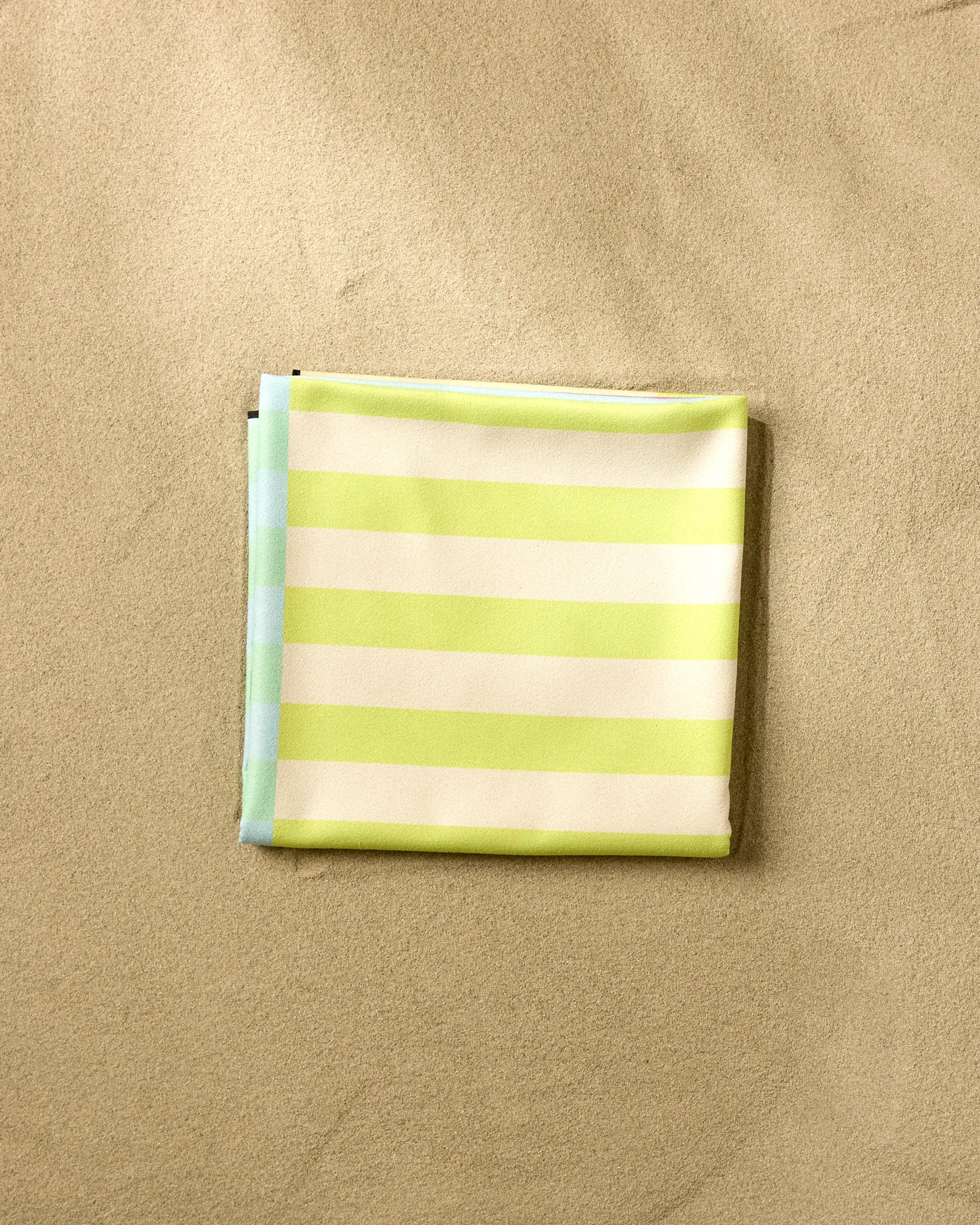 The GEOMETRY Neon Nights beach blanket, featuring light green and white micro-loop stripes with a touch of blue, is folded on the sand.
