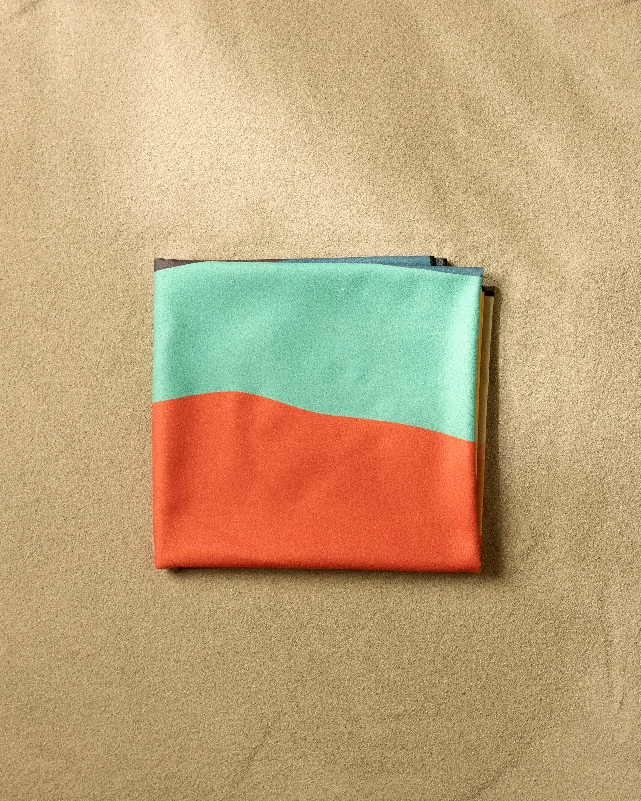 The GEOMETRY Over The Hill folded fabric pouch, featuring a teal and orange color block design, rests on smooth light brown sand—ideal for stashing your quick-drying, sand-repelling beach blanket.