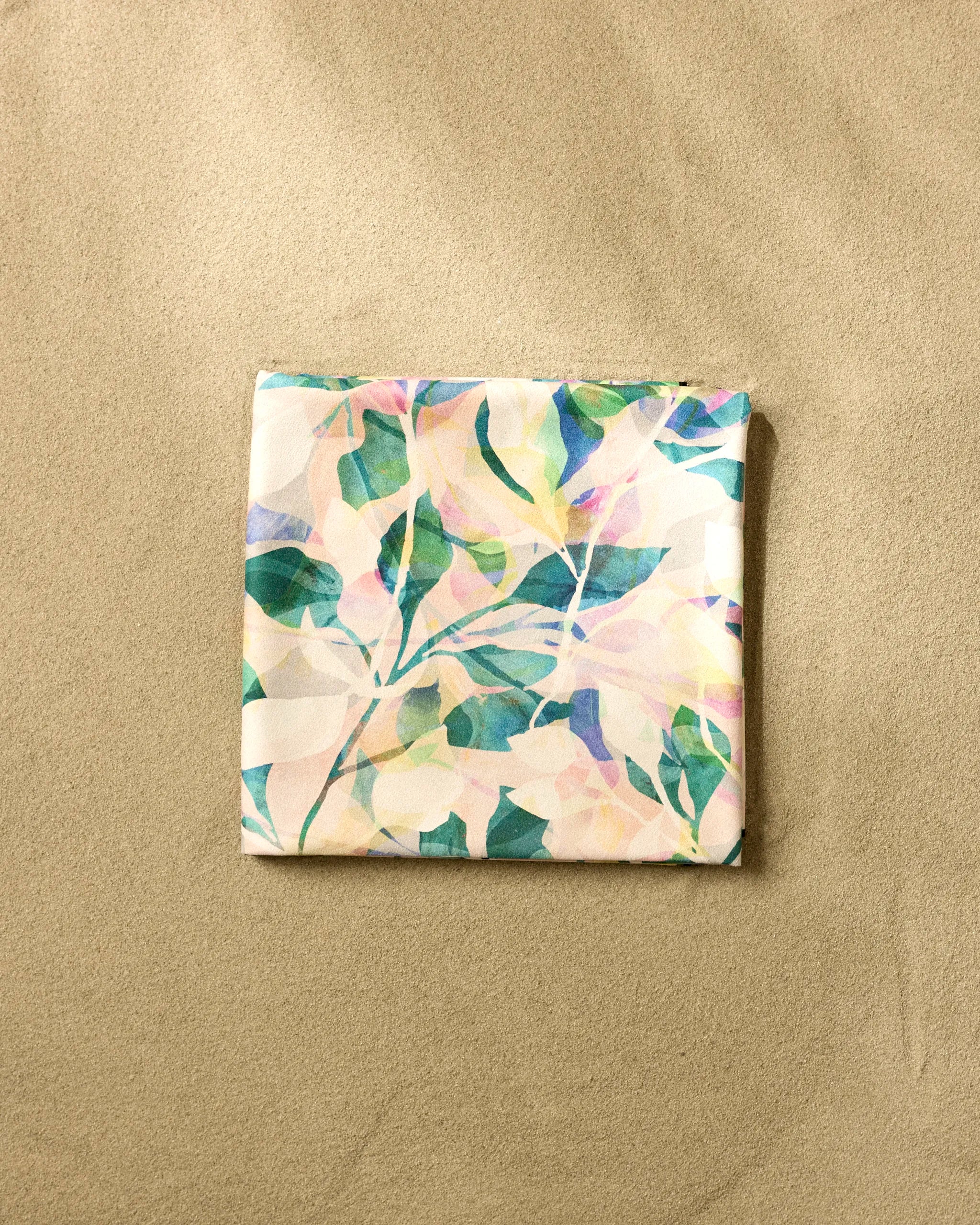 A square, folded Rachel Parker Painted Foliage Micro-loop Beach Blanket featuring a pastel abstract leaf design rests on smooth beige sand.