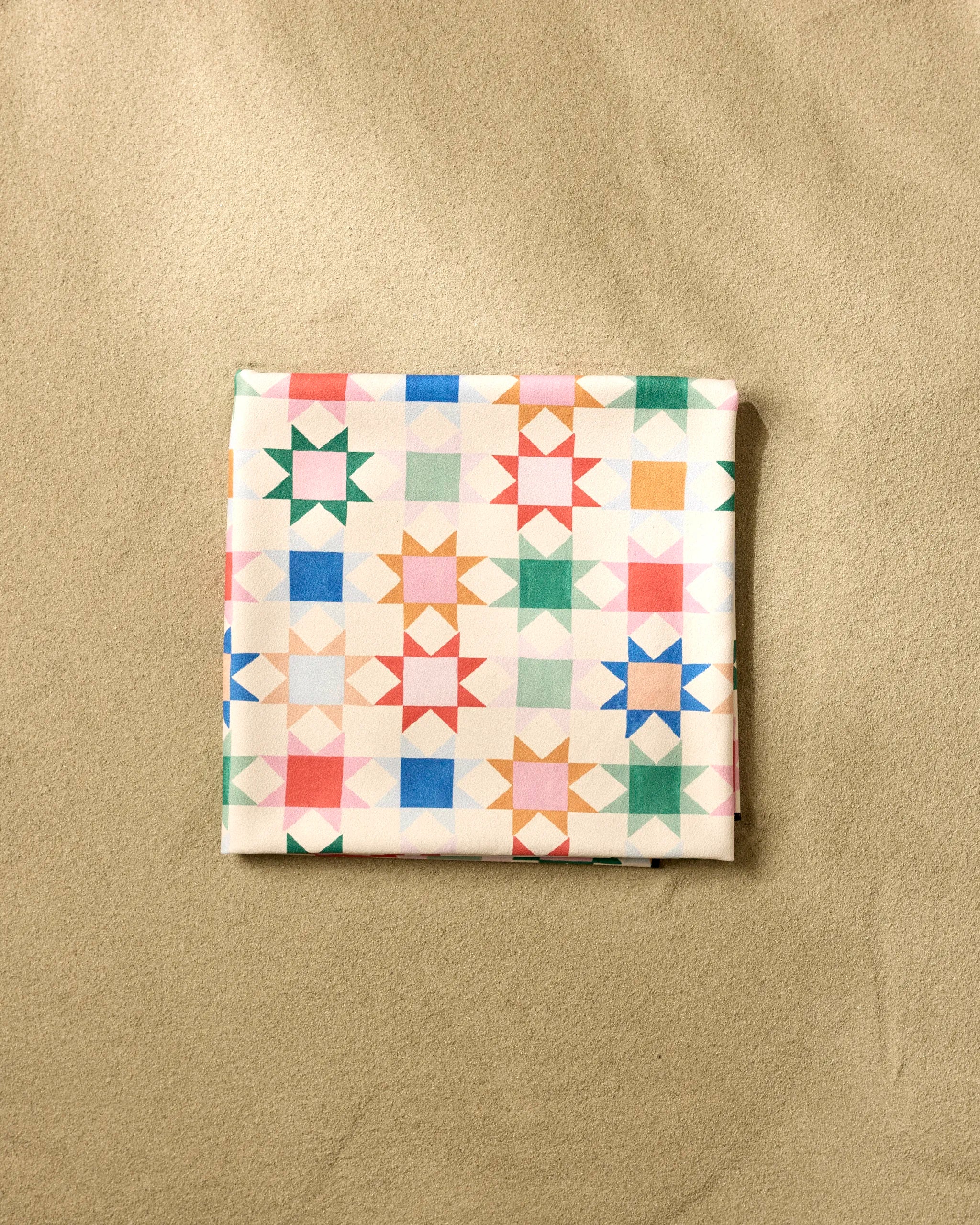 The Patchwork Picnic beach blanket by Brittany Jepsen features a colorful geometric star design and quick-drying fabric, displayed here on a beige textured surface.