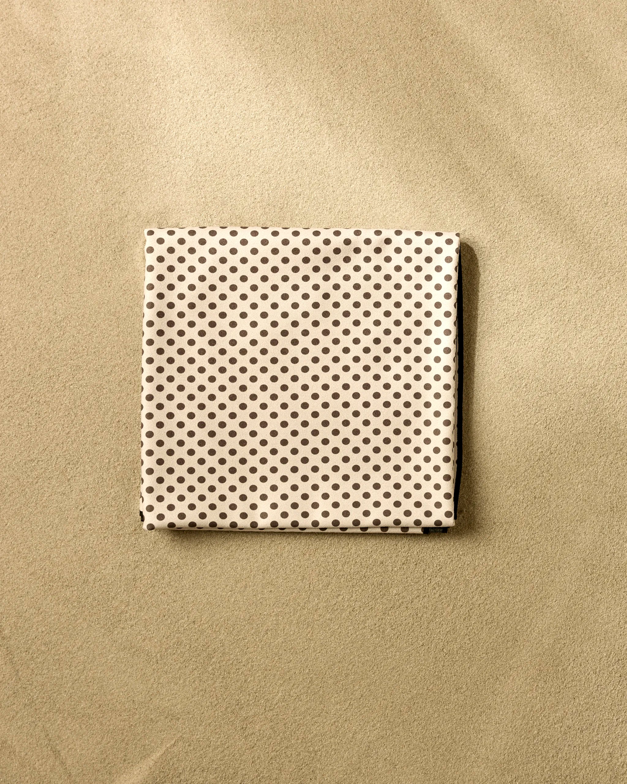 A neatly folded beige cloth with small dark polka dots, the Pebble Beach Blanket by Jenny Keller rests on a light tan surface, blending quick-drying convenience with modern style for your beach days.