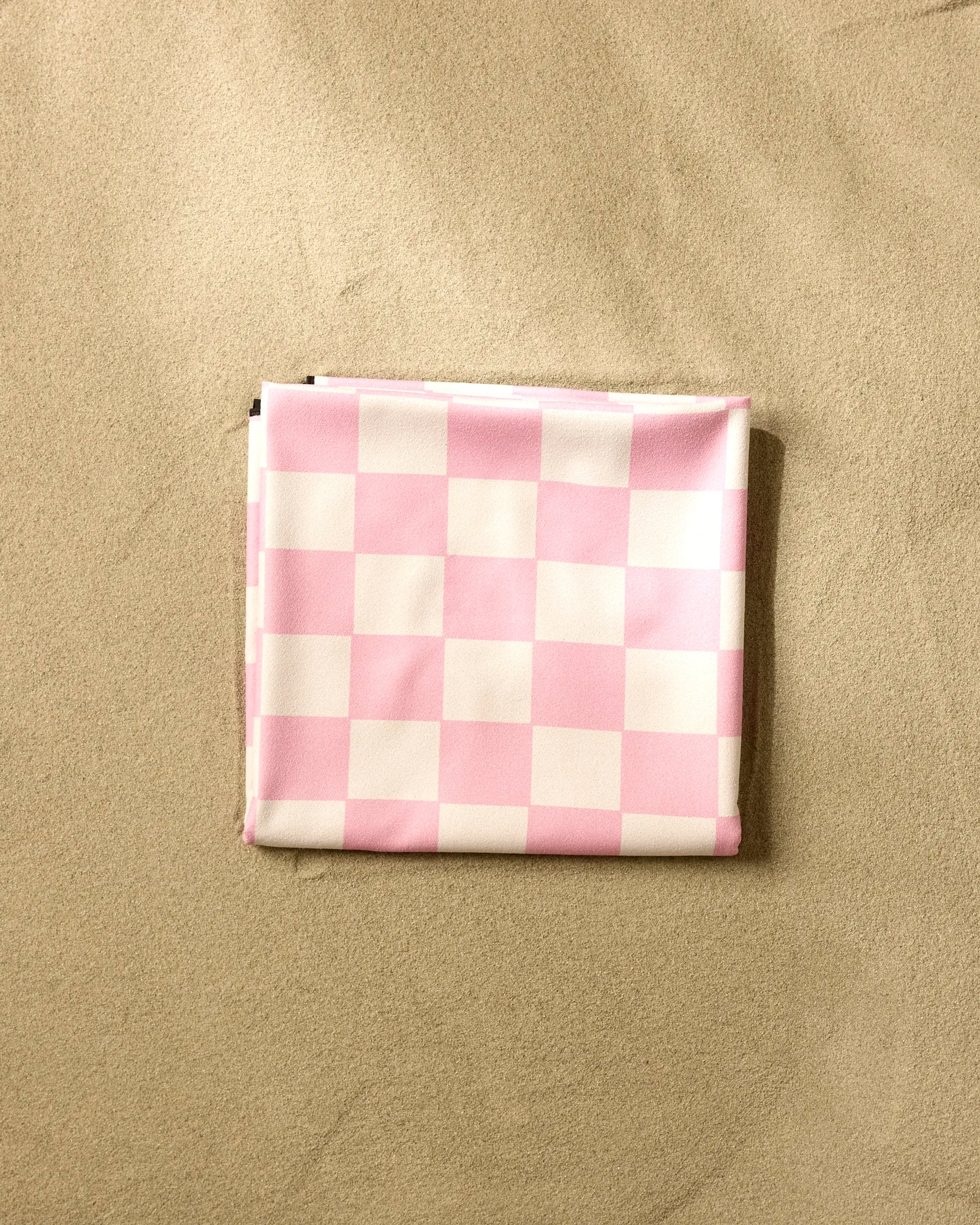 The GEOMETRY Pink Checkers beach blanket, crafted from recycled materials, is neatly folded on light brown sand.