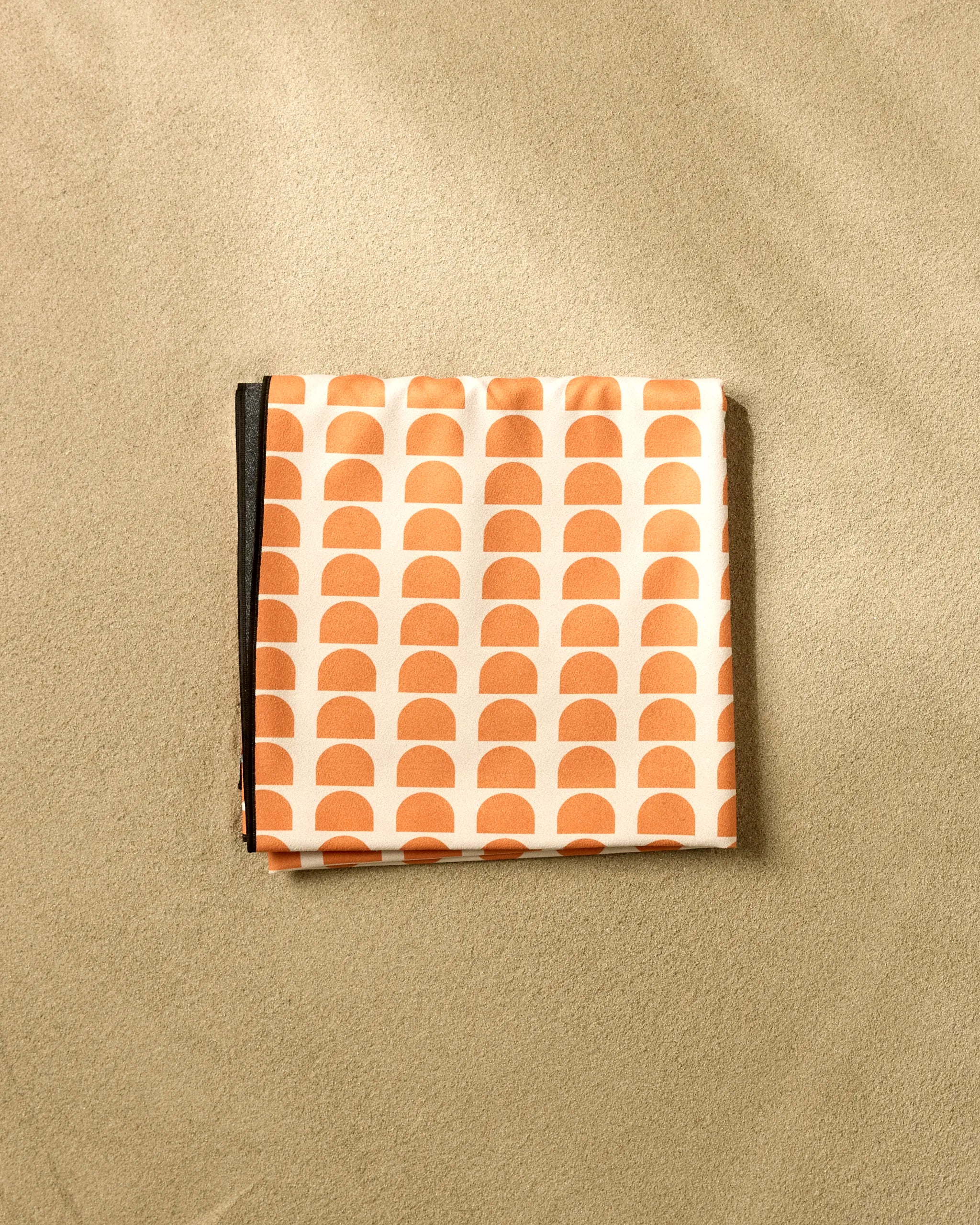 A folded Happy Hemera Pink Illusion Beach Blanket with an orange and white semi-circle pattern rests on sandy beige. The quick-drying blanket features geometric half-circle rows for a bold, modern look.